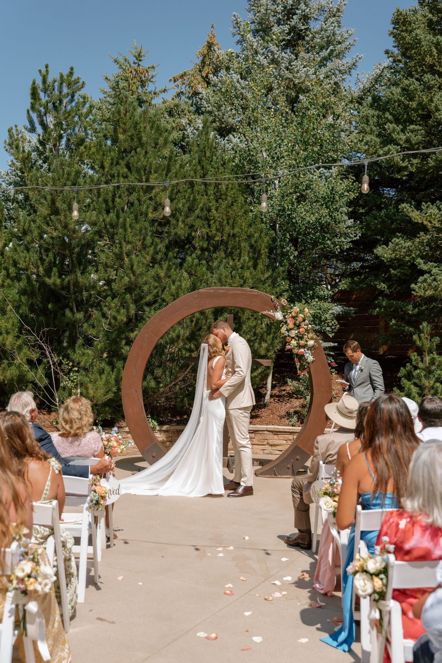Outdoor wedding ceremony with curved wooden arch and white chairs on sandy ground surrounded by pine trees.