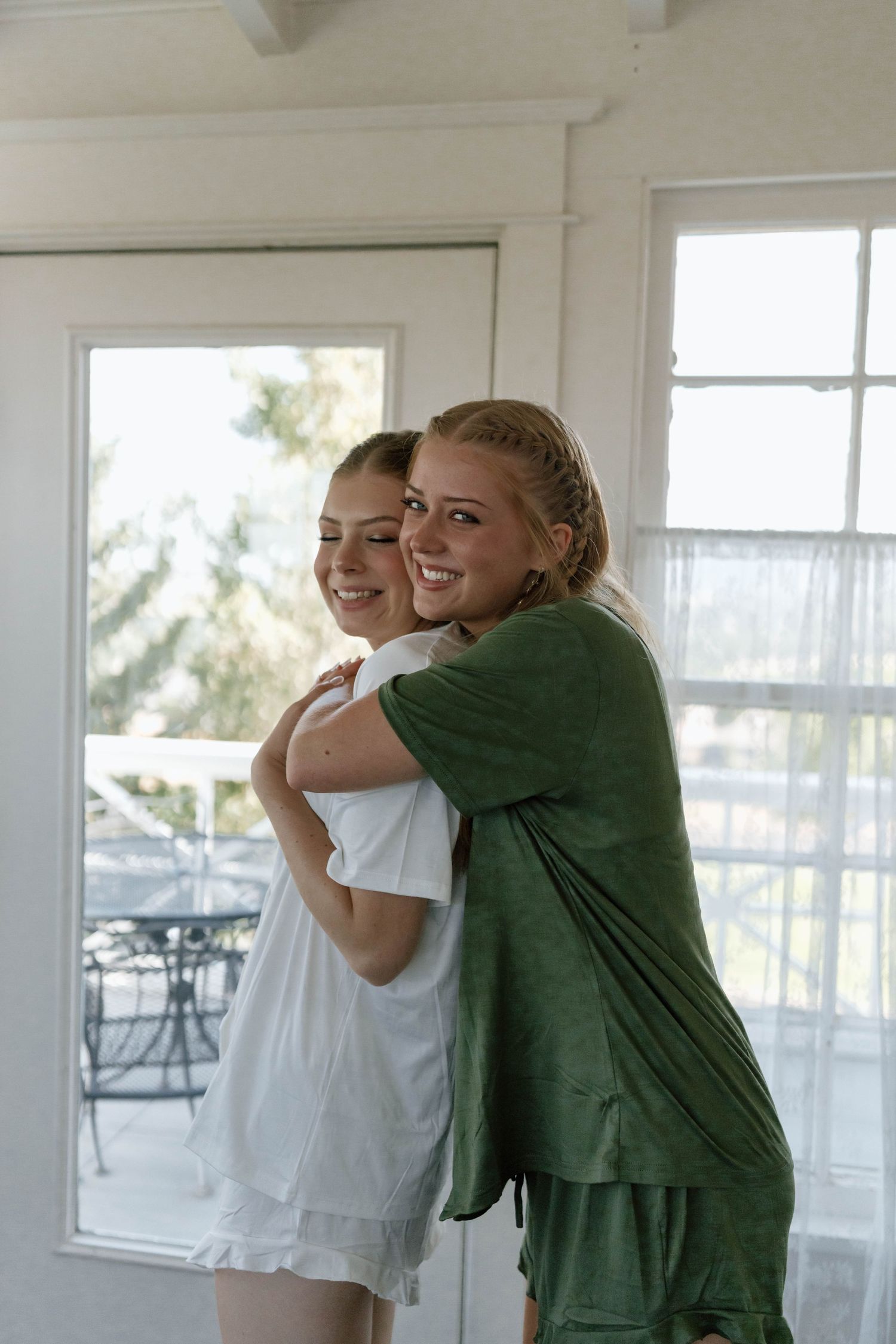 Two people in white and green outfits embrace in a bright sunlit room with large windows.