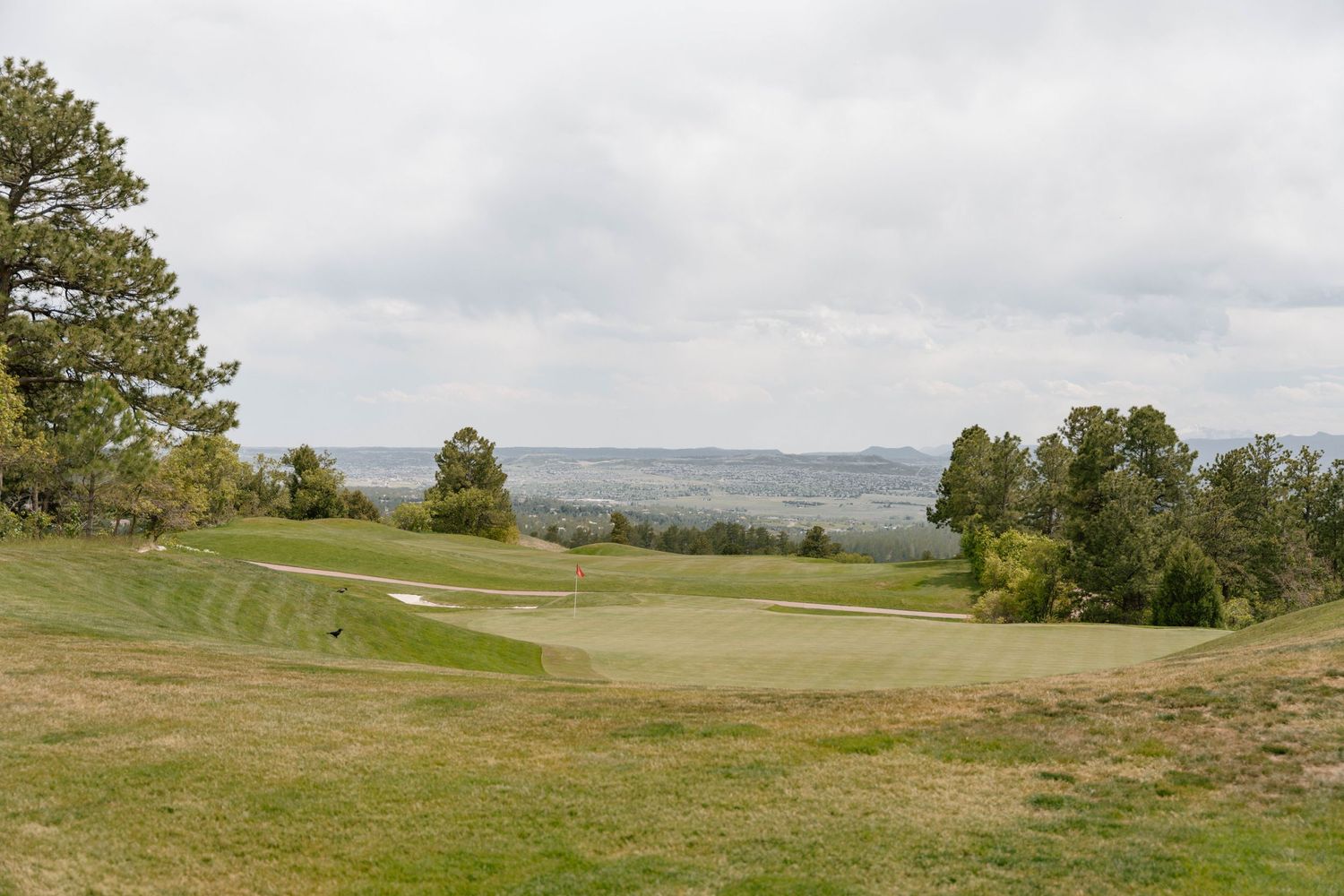 A scenic golf course green surrounded by rolling hills and trees under cloudy skies.
