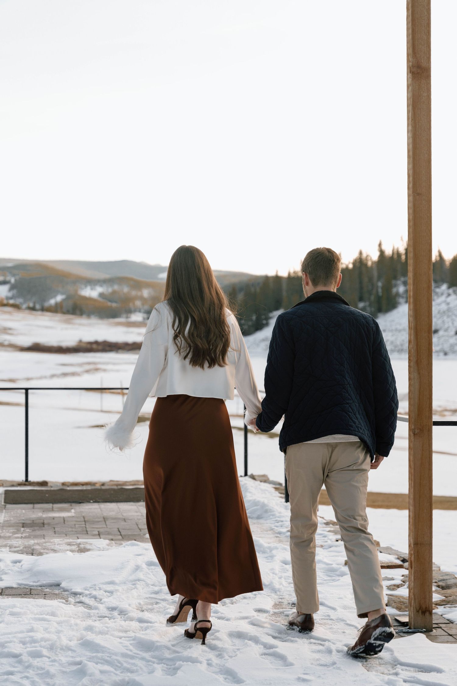 Couple walking on snowy deck with scenic mountain views in winter setting.