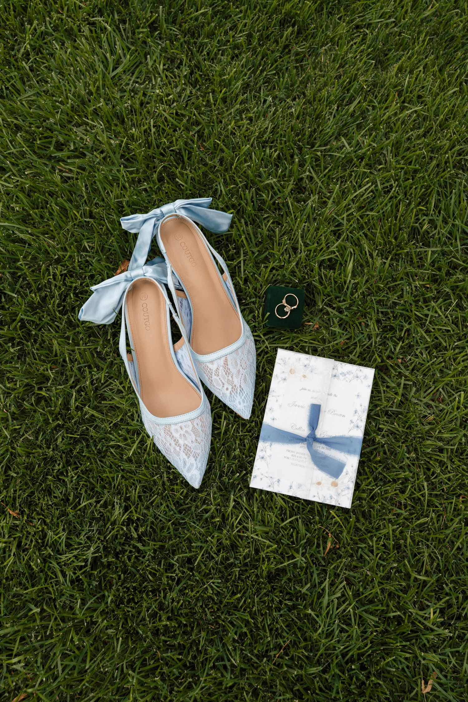 Elegant blue lace bridal heels with satin ribbons on green grass beside wedding rings and floral invitation.