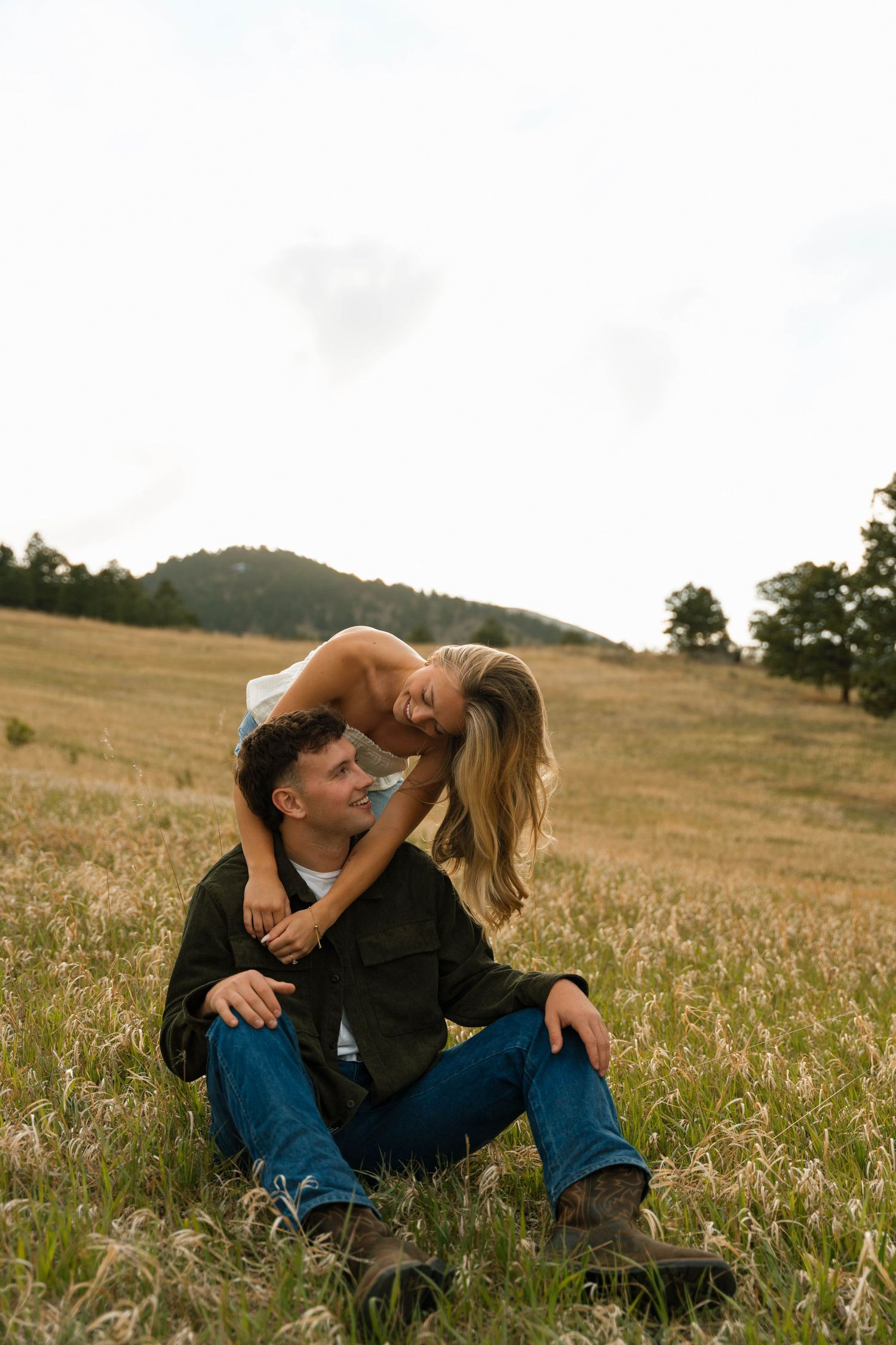 A couple sits in a golden meadow, the woman leaning over the man from behind, both smiling and laughing together.