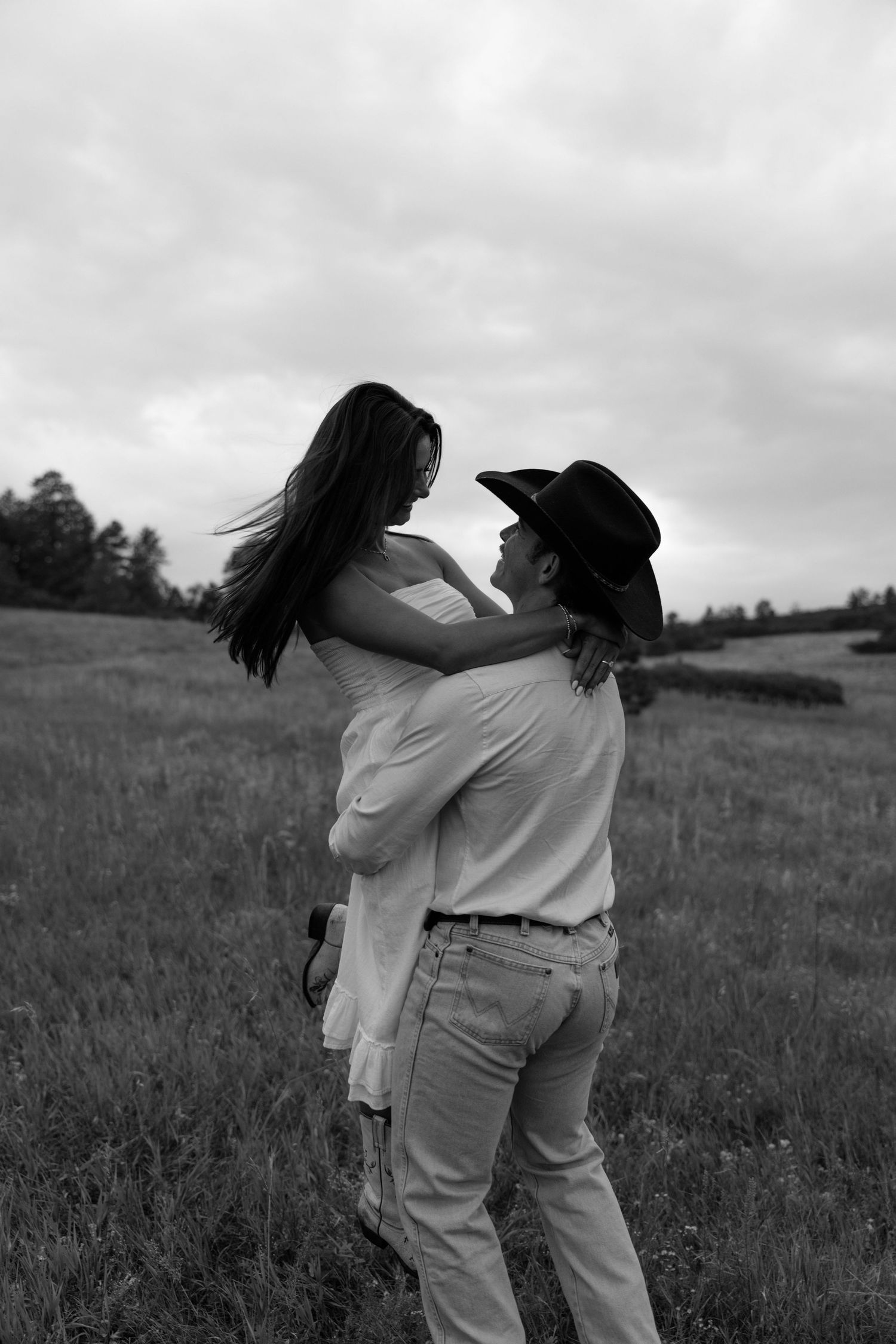 A couple shares a playful moment in a field under cloudy skies in black and white.
