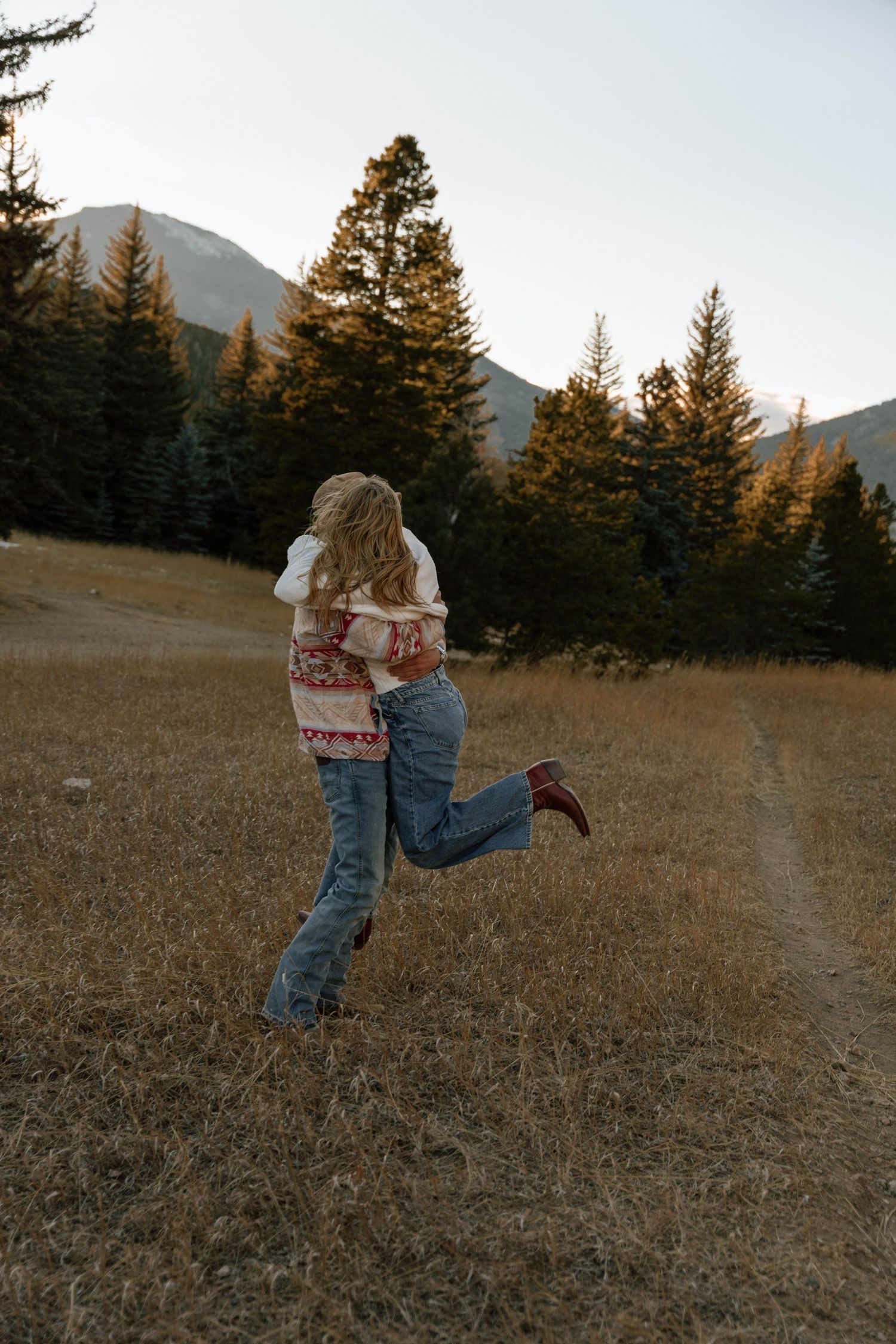 Playful couple dancing in a field at sunset with mountains and evergreen trees in the background.