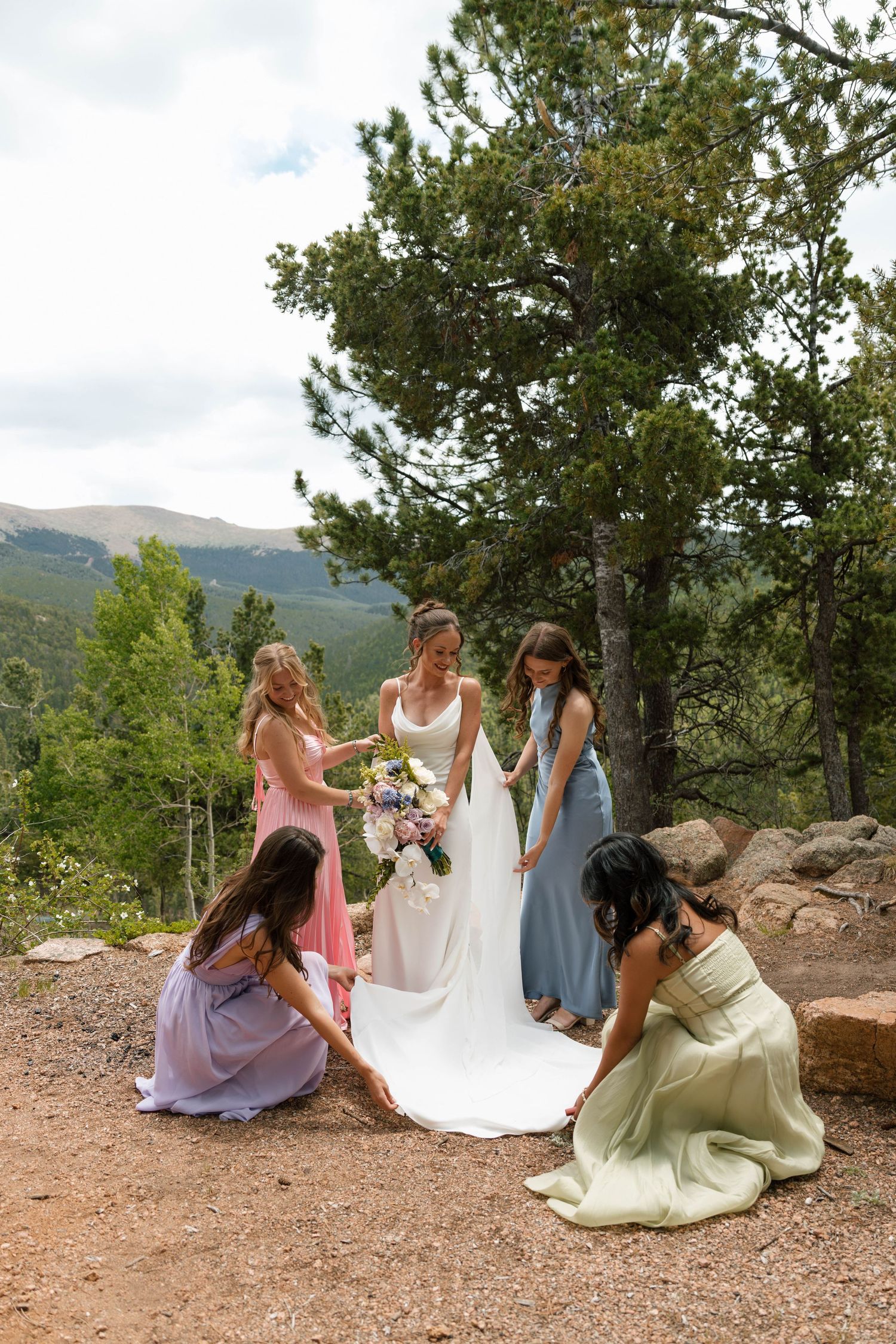 Bride in white gown holds bouquet as bridesmaids in colorful dresses arrange her train on a rocky mountain overlook.