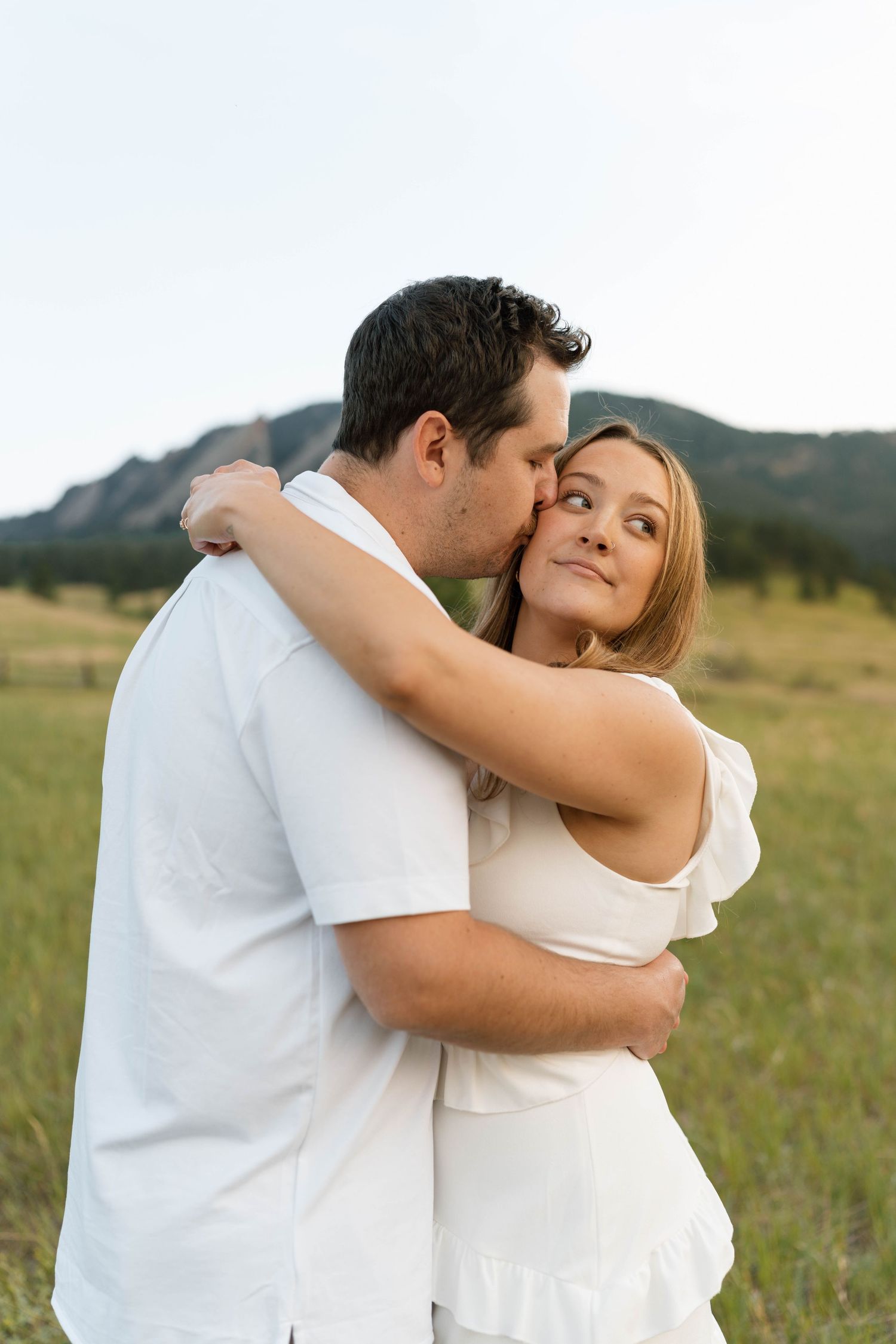 Couple embracing in white outfits in a green mountain meadow, woman smiling as man kisses her cheek.