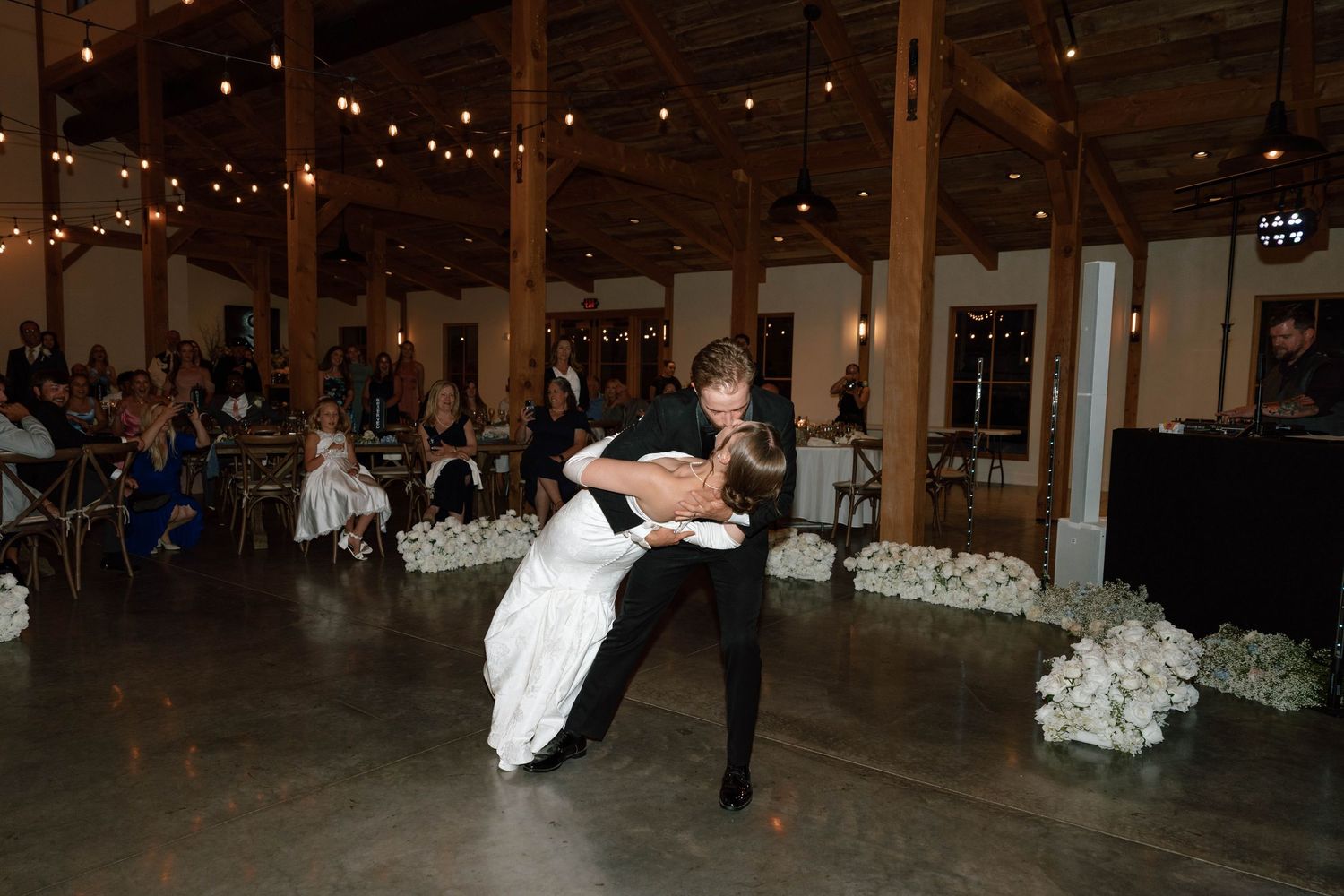 Groom dips bride during romantic first dance at elegant rustic wedding reception venue.