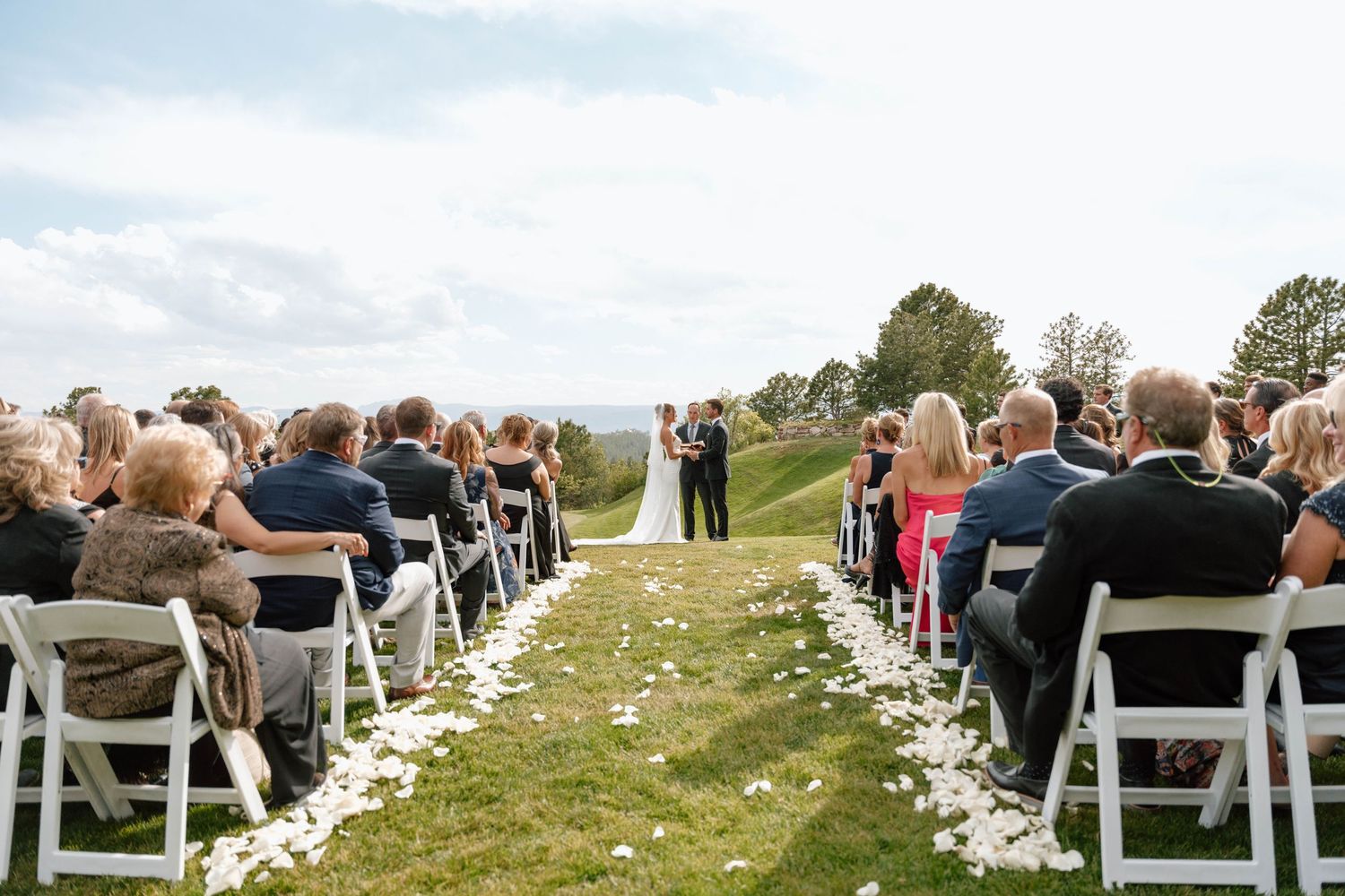 Scenic mountain wedding ceremony with guests seated in white chairs along petal-lined aisle.