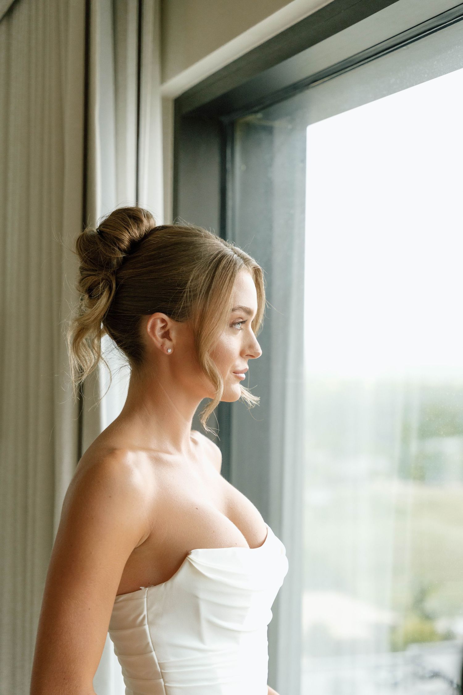 A bride in a strapless white gown gazes out a large window with natural light streaming in.
