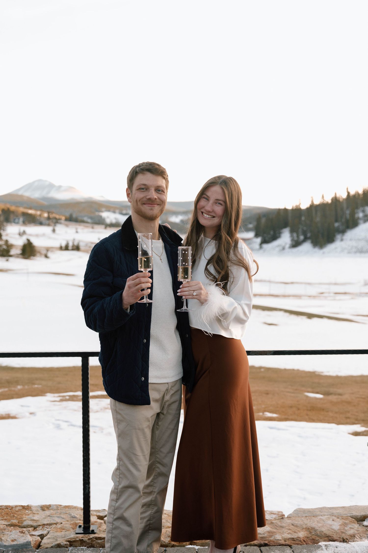 Couple celebrating with drinks on a scenic winter mountain deck.