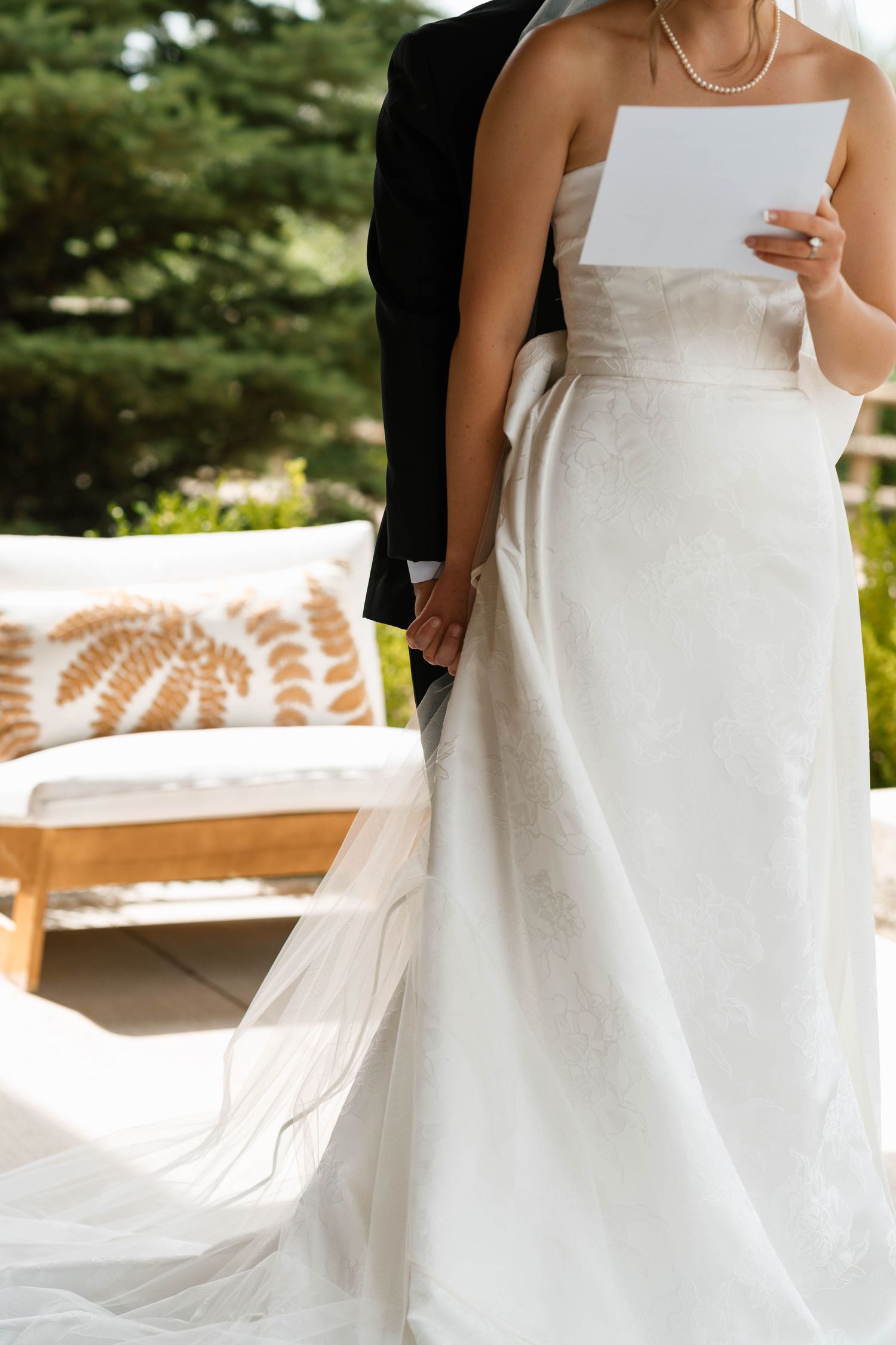 Bride in strapless white gown reads vows while holding groom's hand at outdoor wedding ceremony.