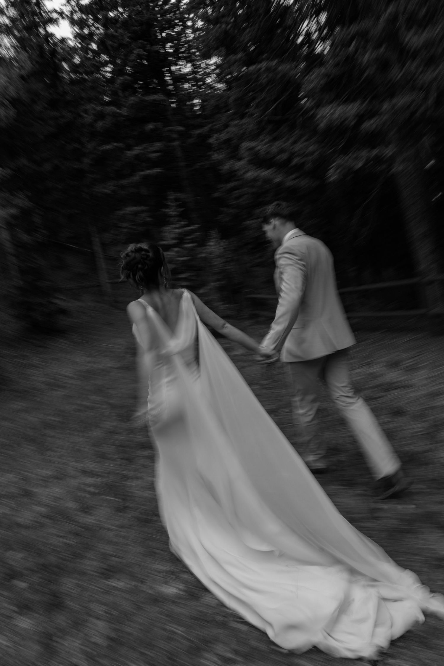 Blurred motion shot of bride and groom holding hands walking through a dark forest path.