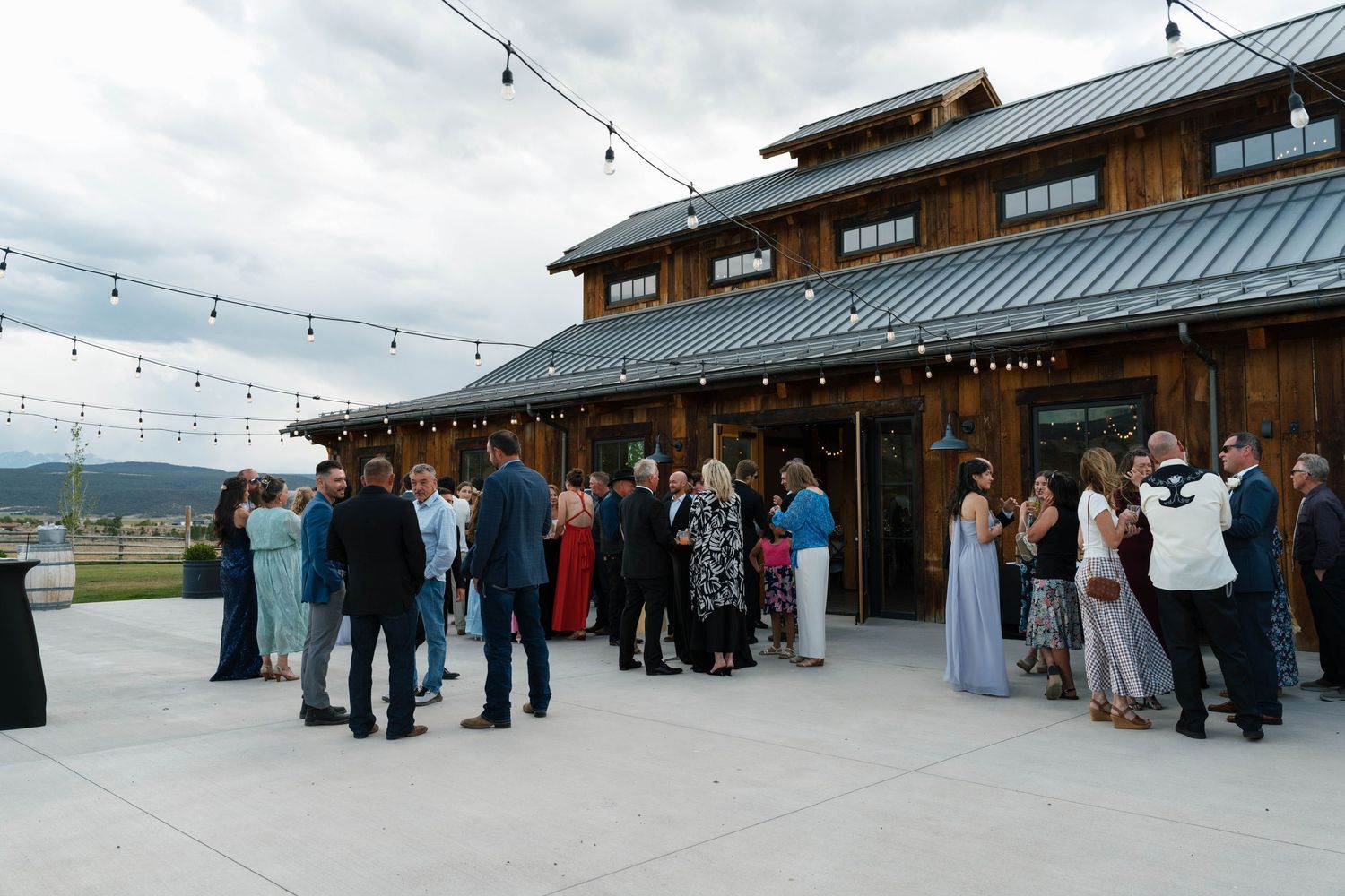 Wedding guests mingle on outdoor patio of rustic barn venue with string lights.