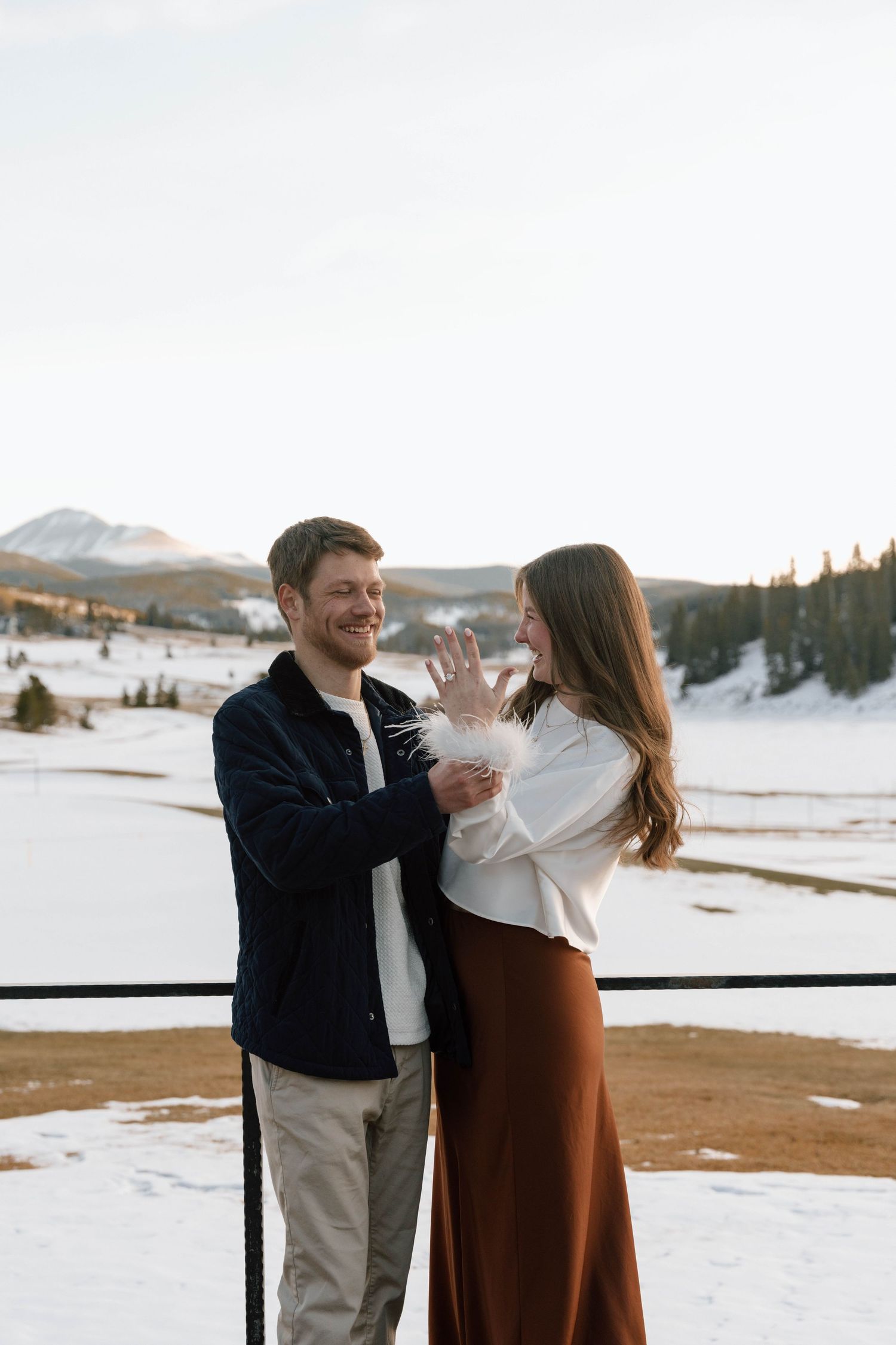 Happy couple celebrating engagement on mountain deck with snow-covered scenery.