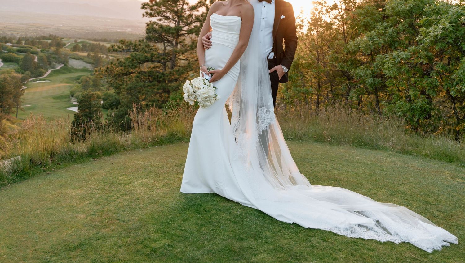 Wedding couple photographed from behind with long flowing dress train on scenic hilltop at sunset.
