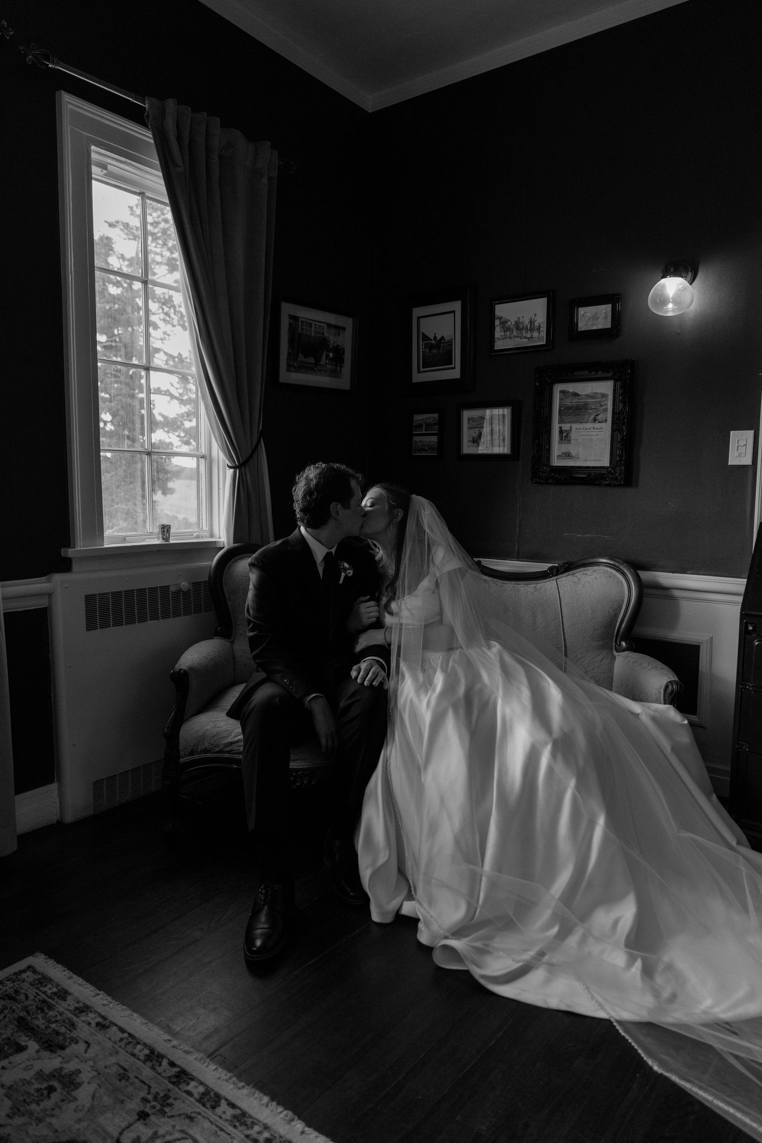 Black and white photo of bride and groom sharing a kiss on a vintage sofa by a window.