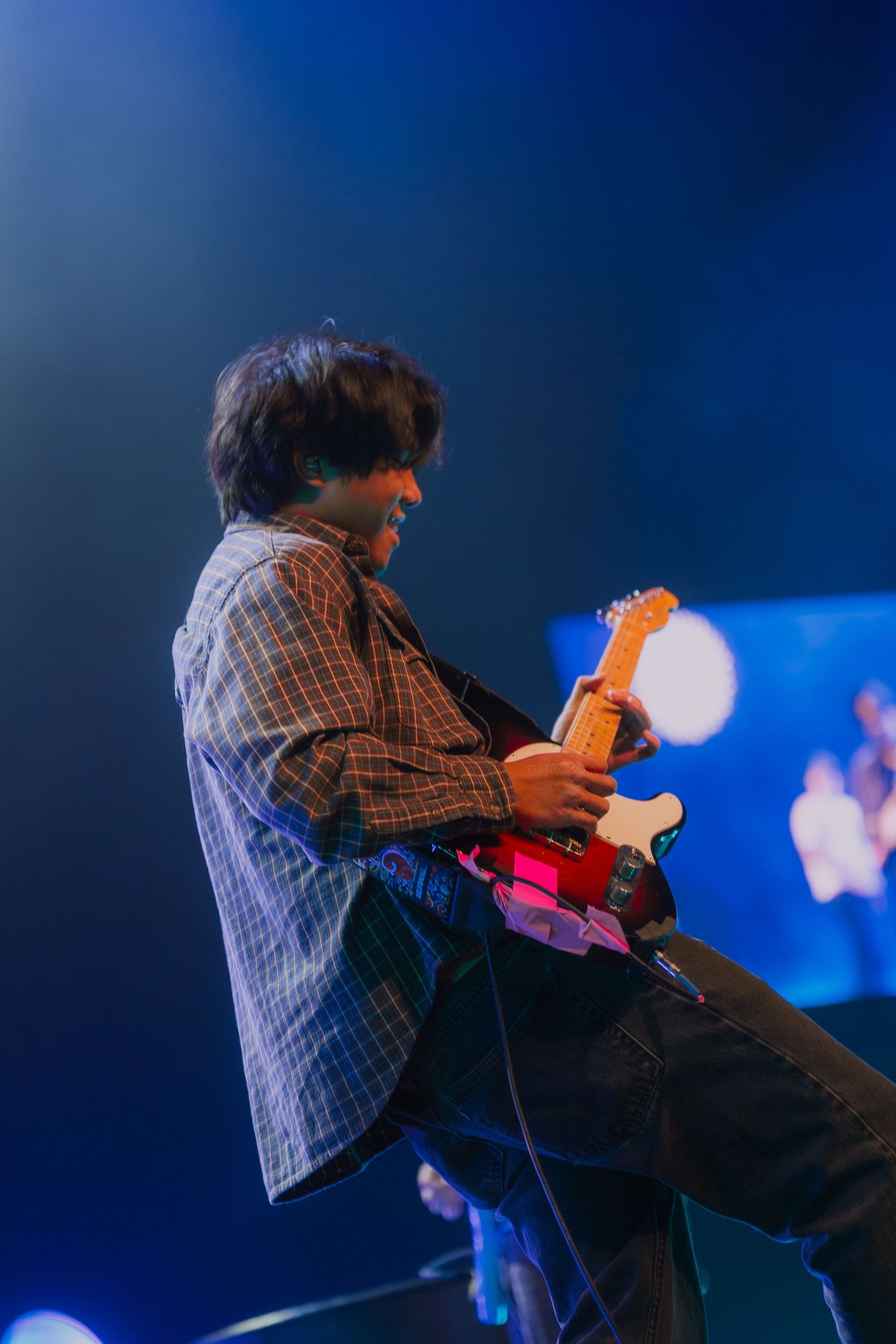 A performer in a plaid shirt plays guitar on stage with dramatic blue lighting and bokeh effects in the background.