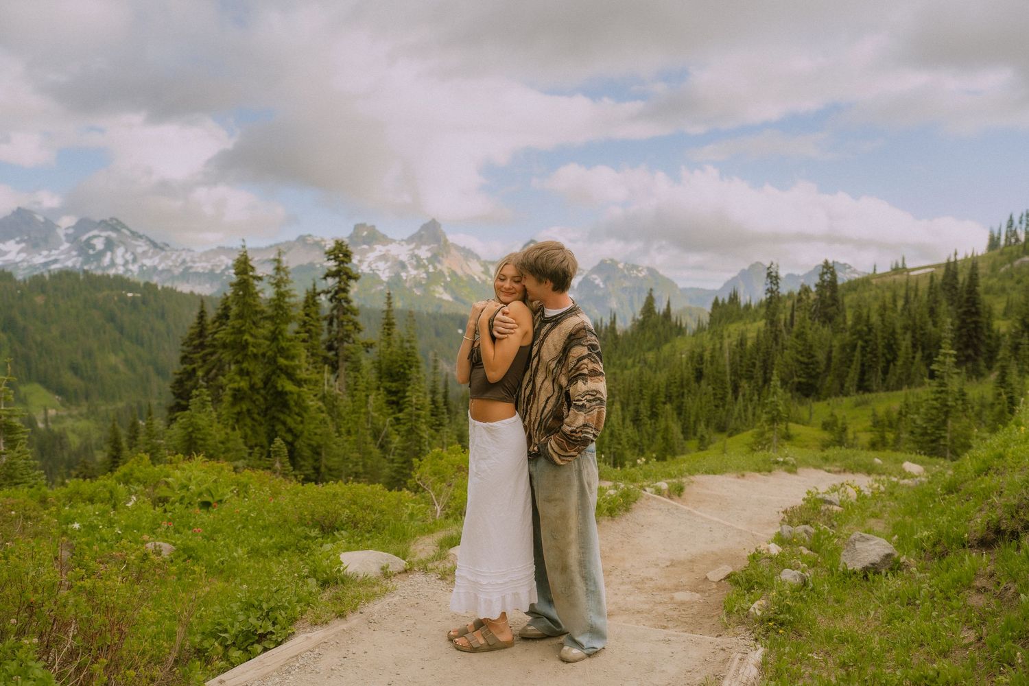 A couple walks along a mountain trail surrounded by evergreen trees and snow-capped peaks in the distance.