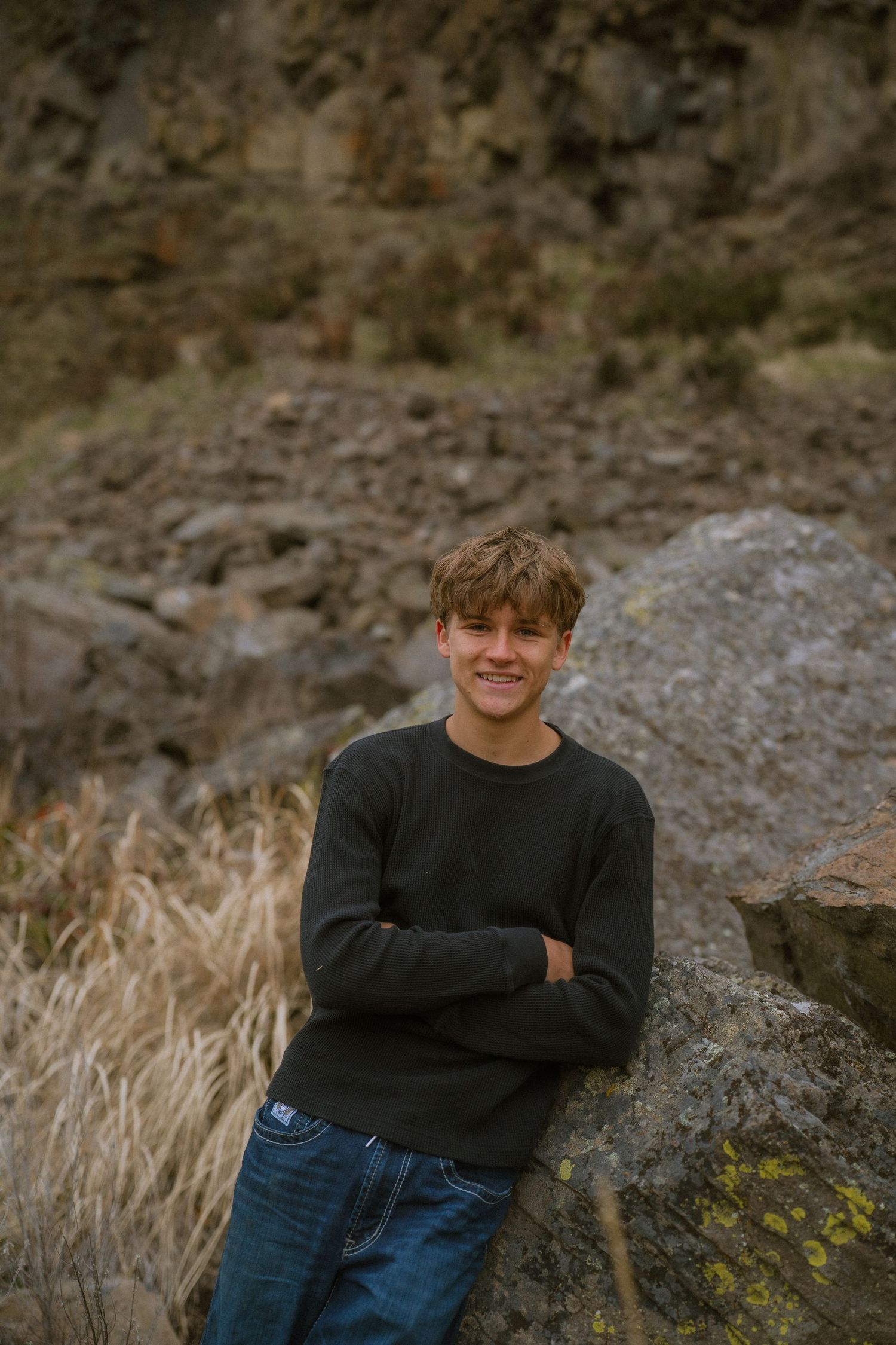 Person in black sweater and jeans poses against rocky outdoor backdrop during golden hour.