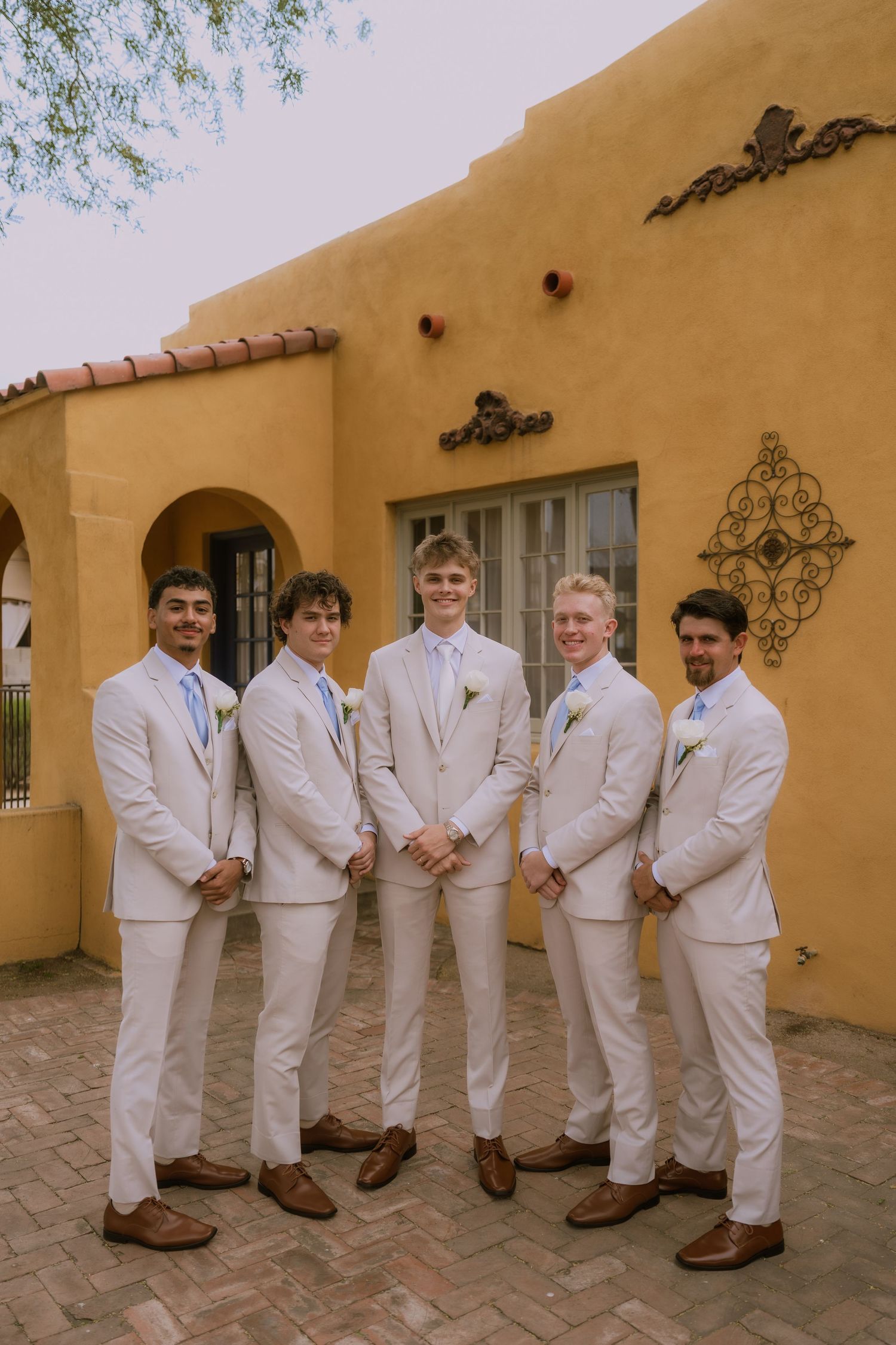 Groomsmen in white suits pose together against a southwestern style yellow adobe wall.