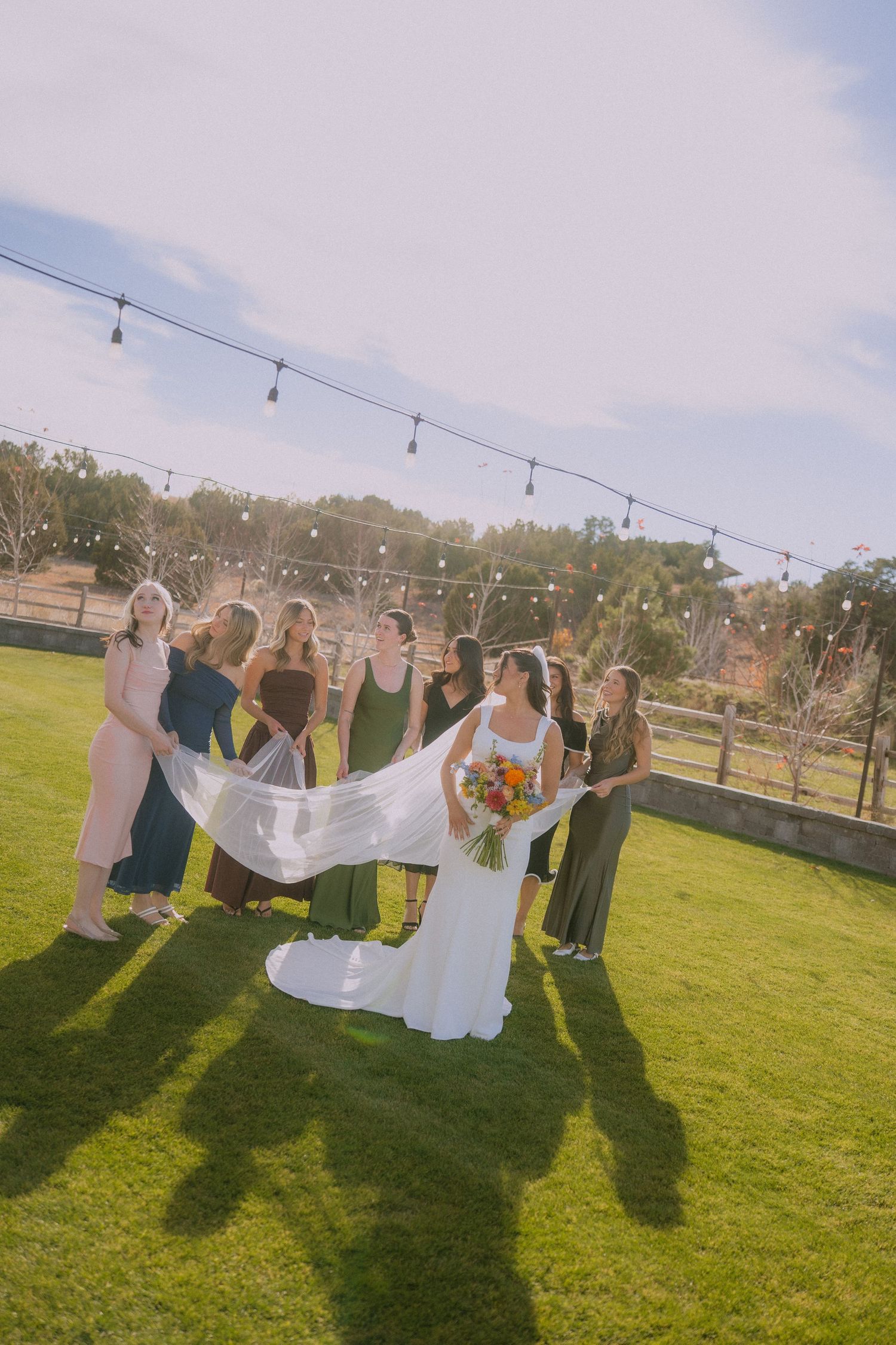 Wedding party members pose with a flowing veil against a sunny outdoor backdrop with string lights.