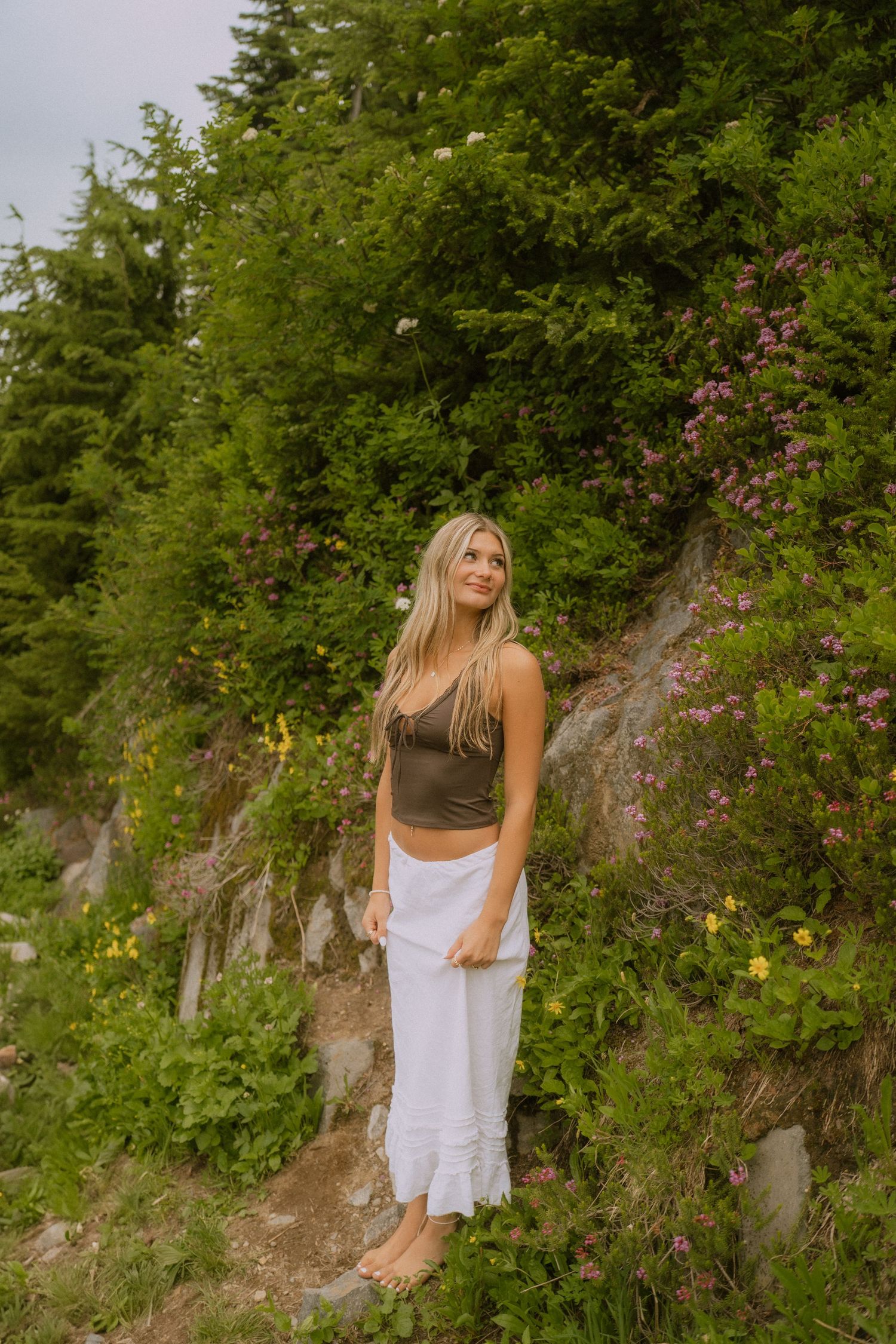 A figure stands on a rocky hillside wearing a brown tank top and white skirt among flowering bushes and greenery.