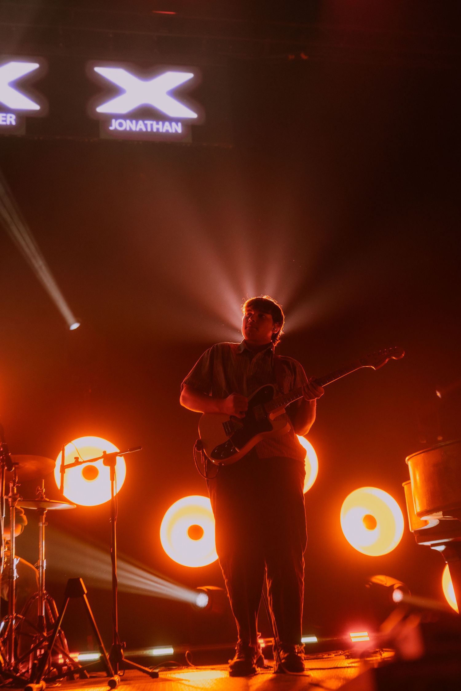 Musicians perform on stage with dramatic orange backlighting and silhouetted figures creating a moody concert atmosphere.