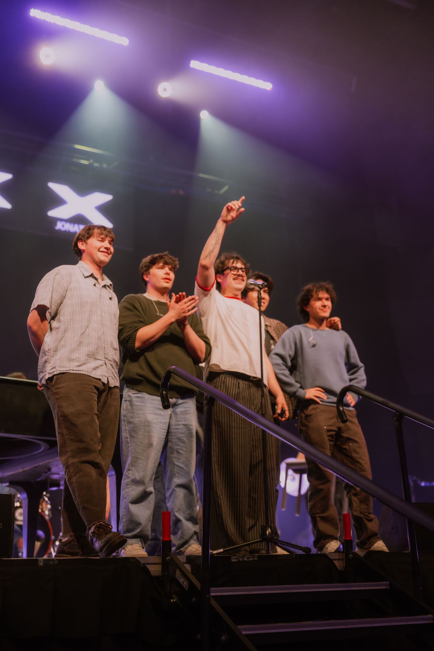 Group of performers stand in formation on stage steps under purple and white theatrical lighting during a show.