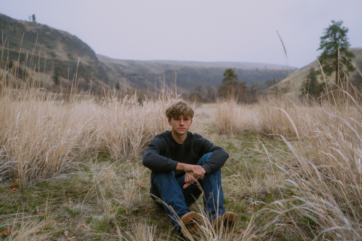 A person sits in tall dry grass on a hillside overlooking a scenic mountain landscape on an overcast day.