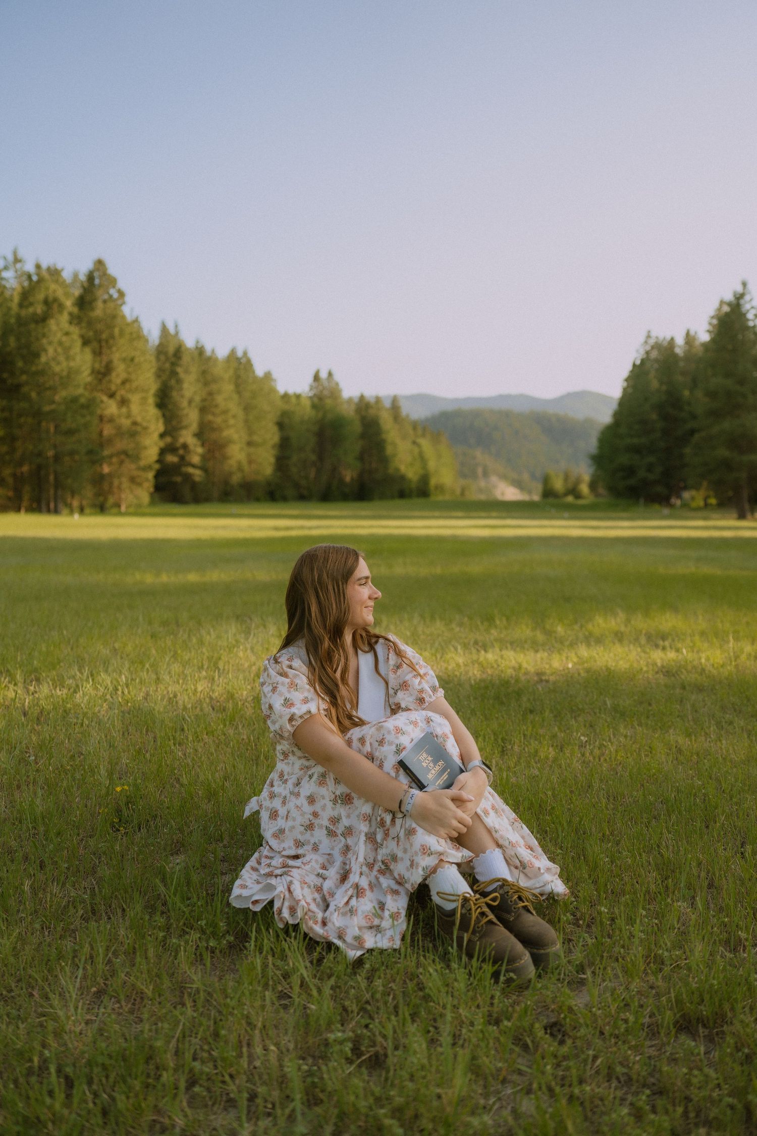 A floral dress-clad figure sits peacefully in a sunlit meadow with mountains and forest in the background.