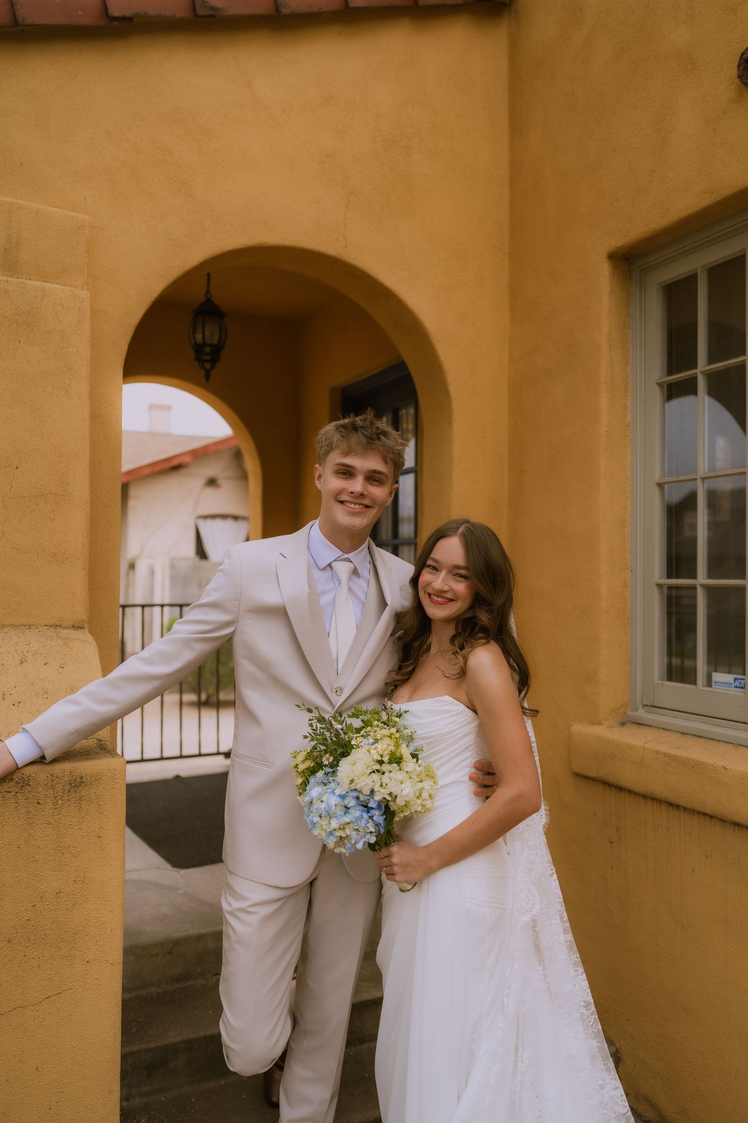 A wedding couple shares candid moments in front of a Spanish-style stucco building with arched doorways.