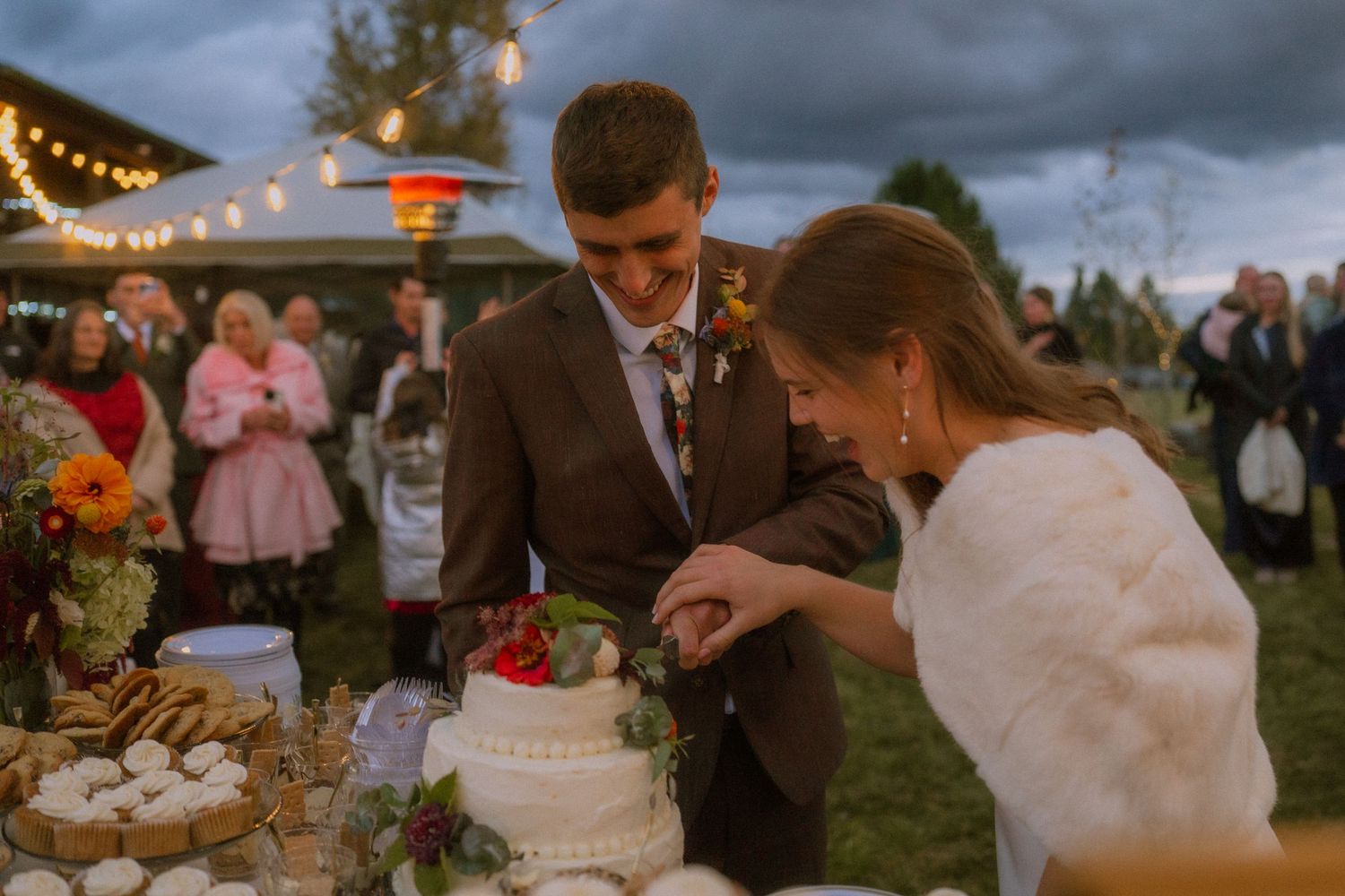 A romantic outdoor wedding reception at dusk with string lights, dancing, and a white tiered cake on display.