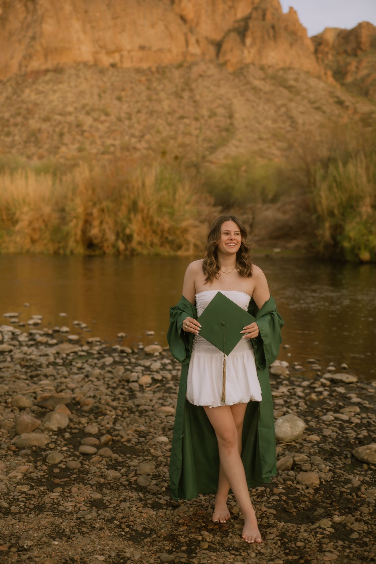 A barefoot woman in a white dress and green robe stands by a rocky riverbank with cliffs in the background at sunset.