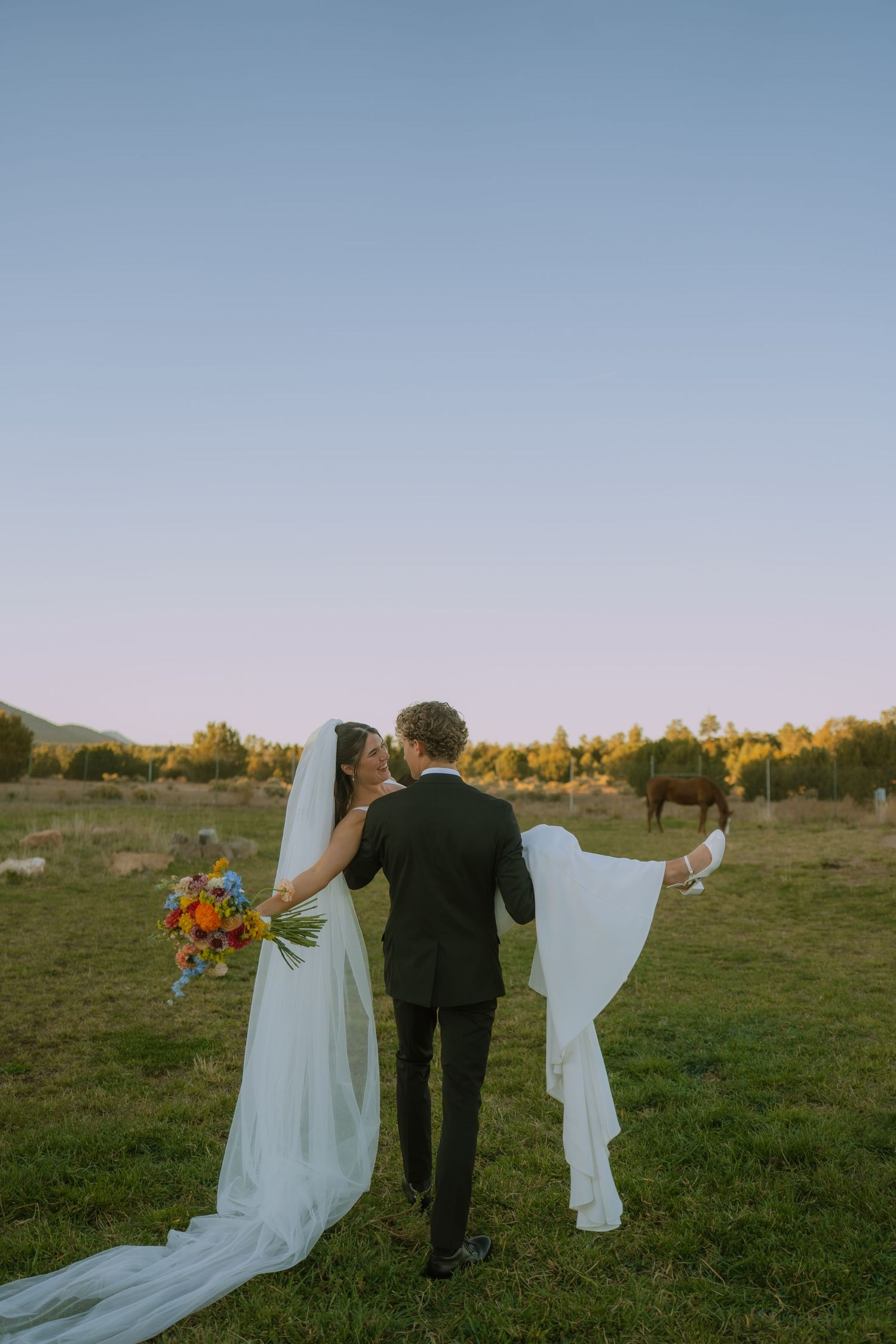A couple in wedding attire walks together across a grassy field at sunset with mountains in the distance.