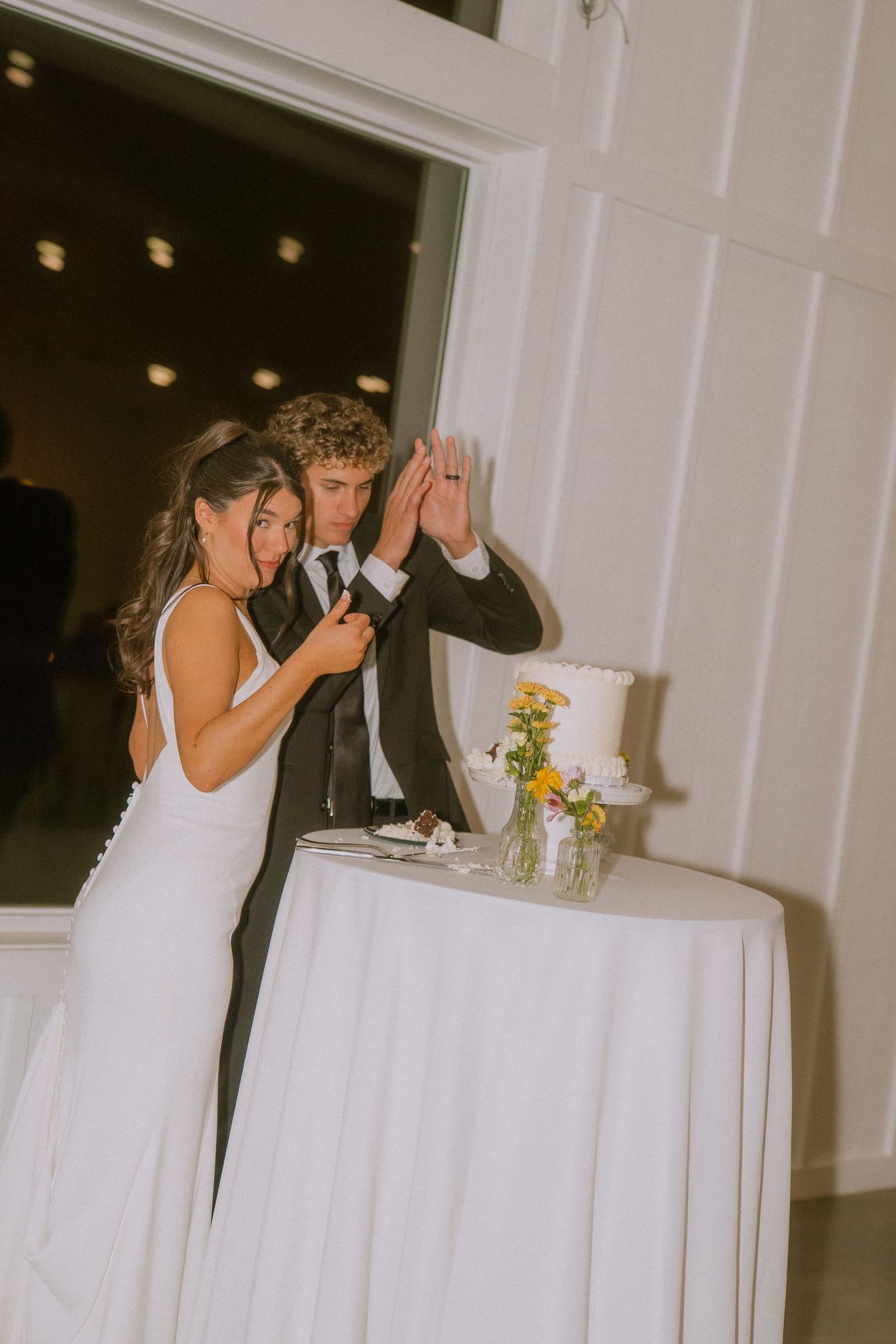 Couple in wedding attire celebrating playfully by cake table indoors.