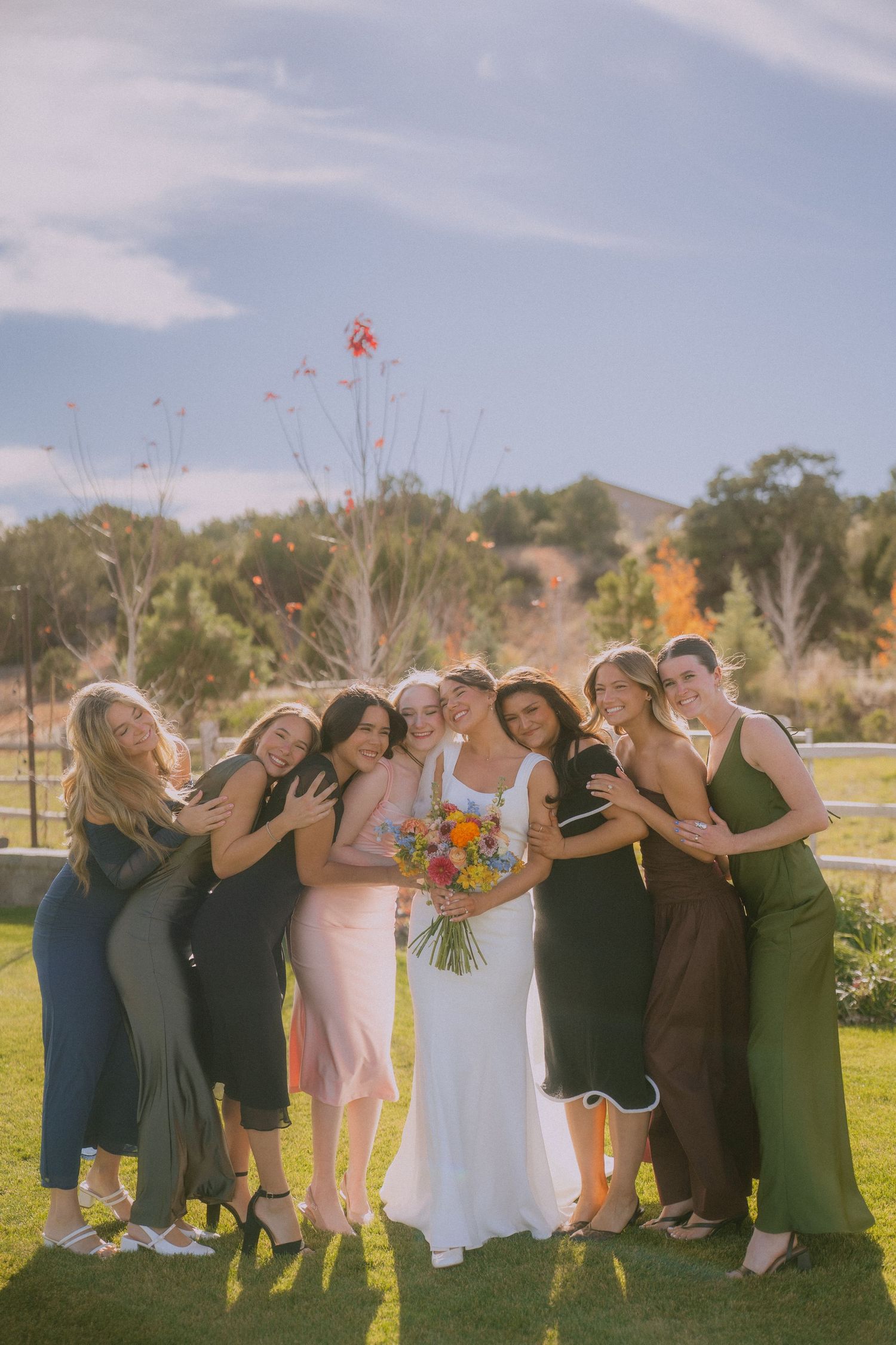 Wedding party group poses playfully in a sunny outdoor field with colorful bouquets.