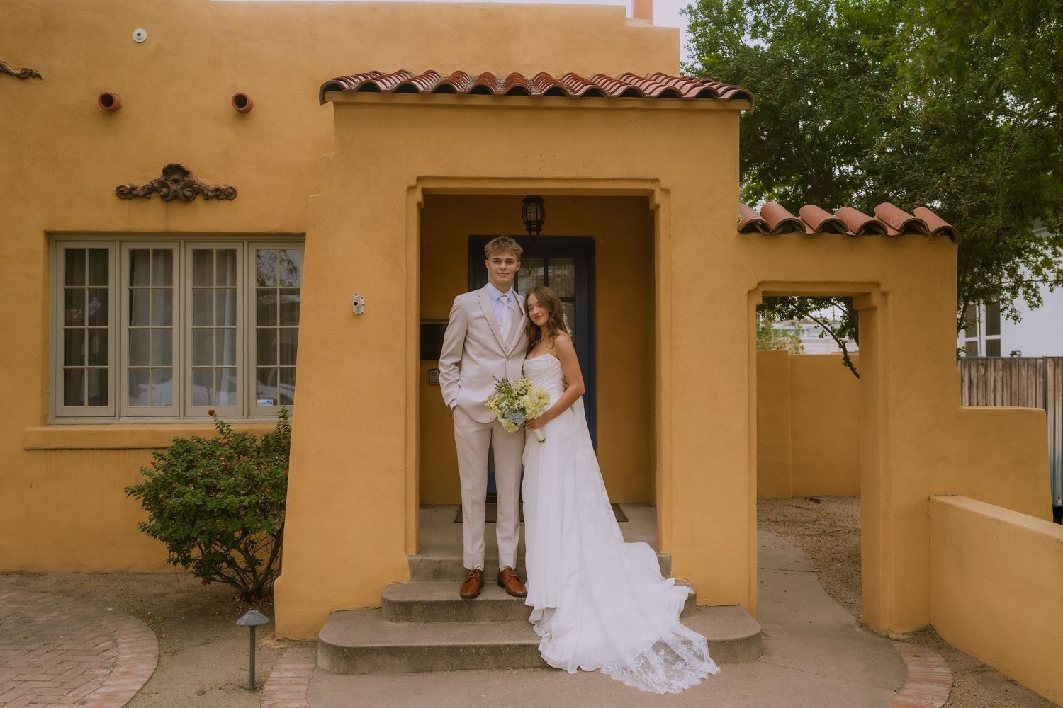 A newlywed couple poses on the steps of a yellow Mediterranean-style house with terracotta roof tiles.