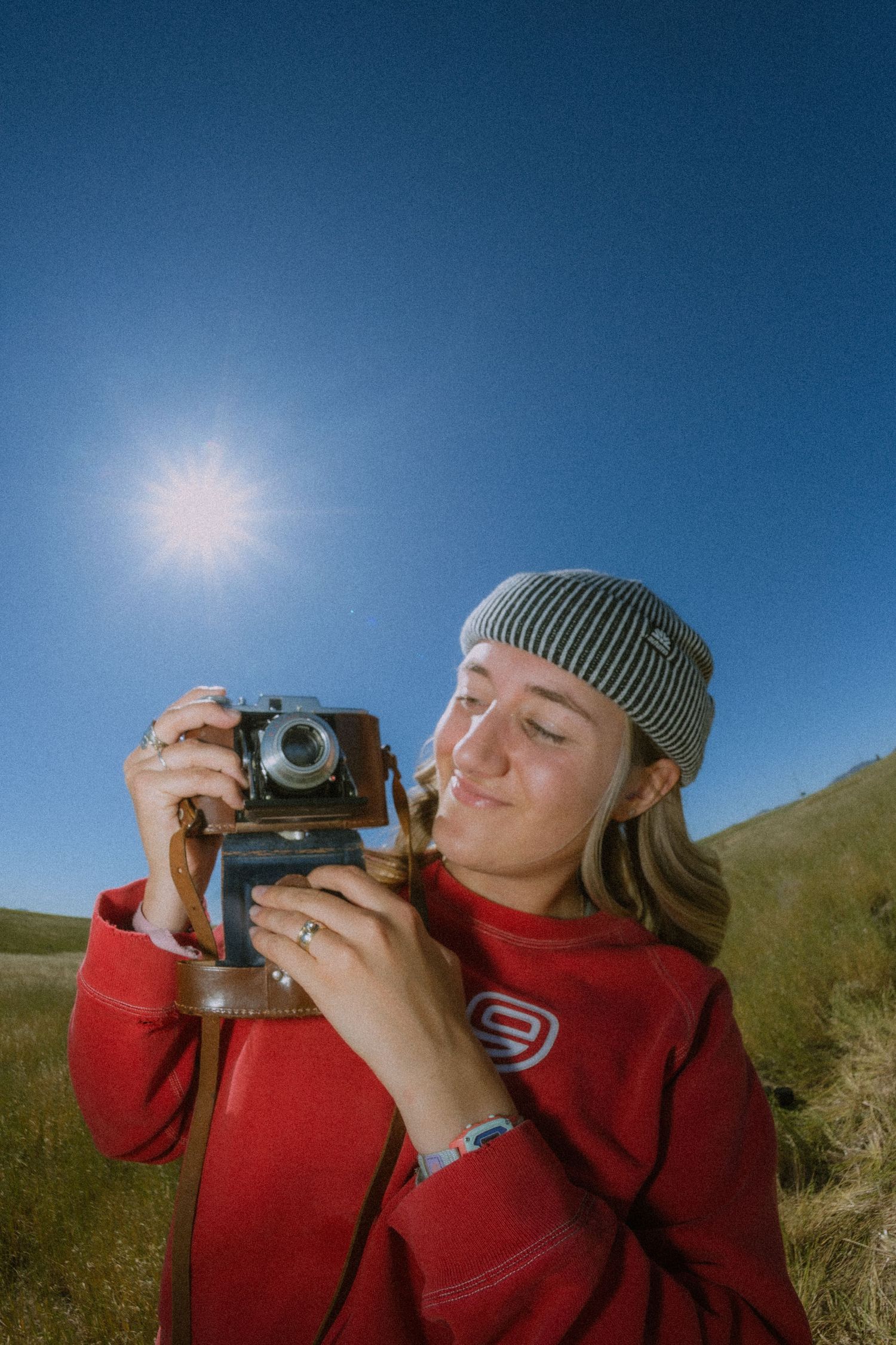 A photographer in a red sweater and gray beanie takes photos with a vintage camera against a blue sky backdrop.