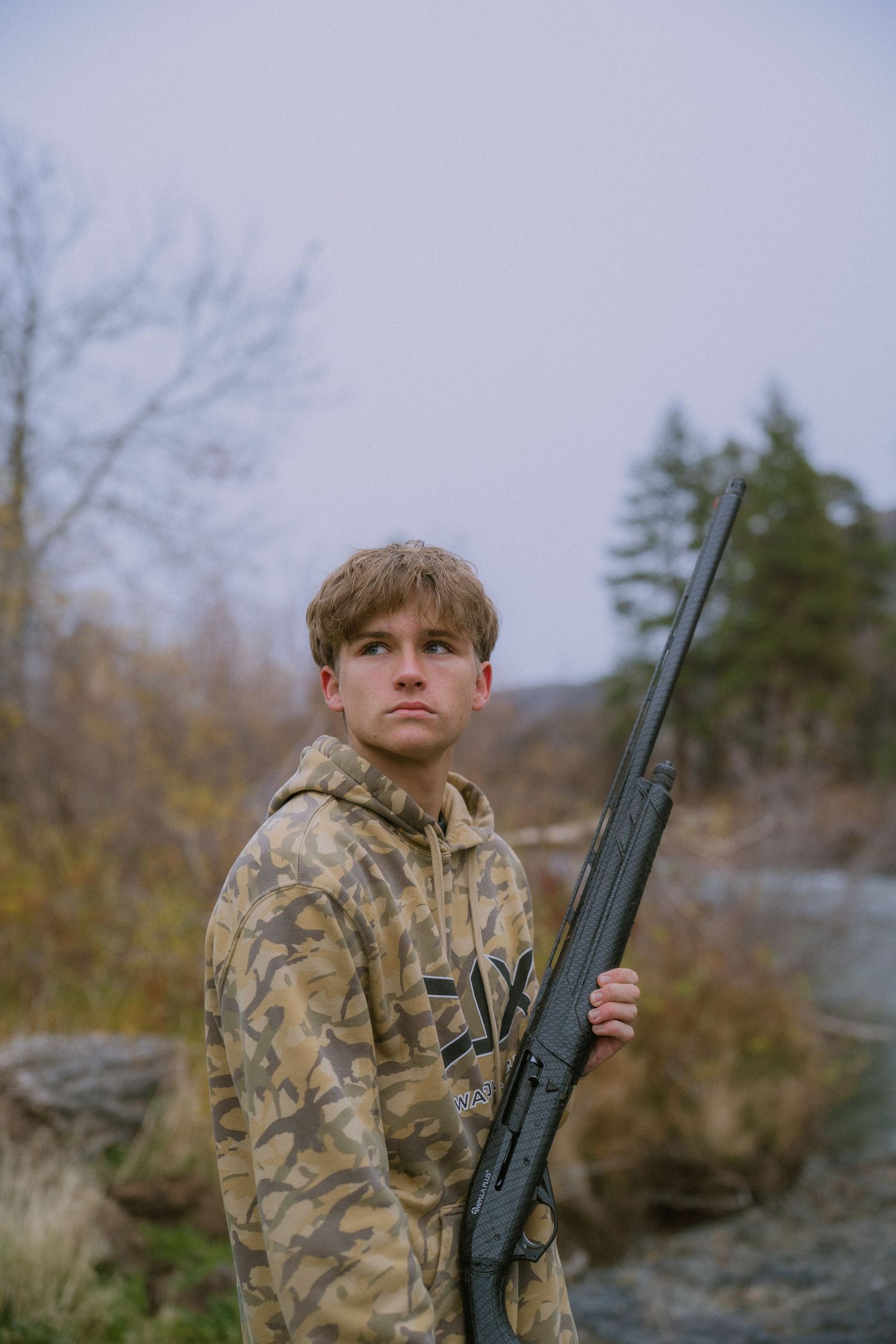 Hunter in camouflage clothing holds a rifle while standing outdoors in an autumn landscape.