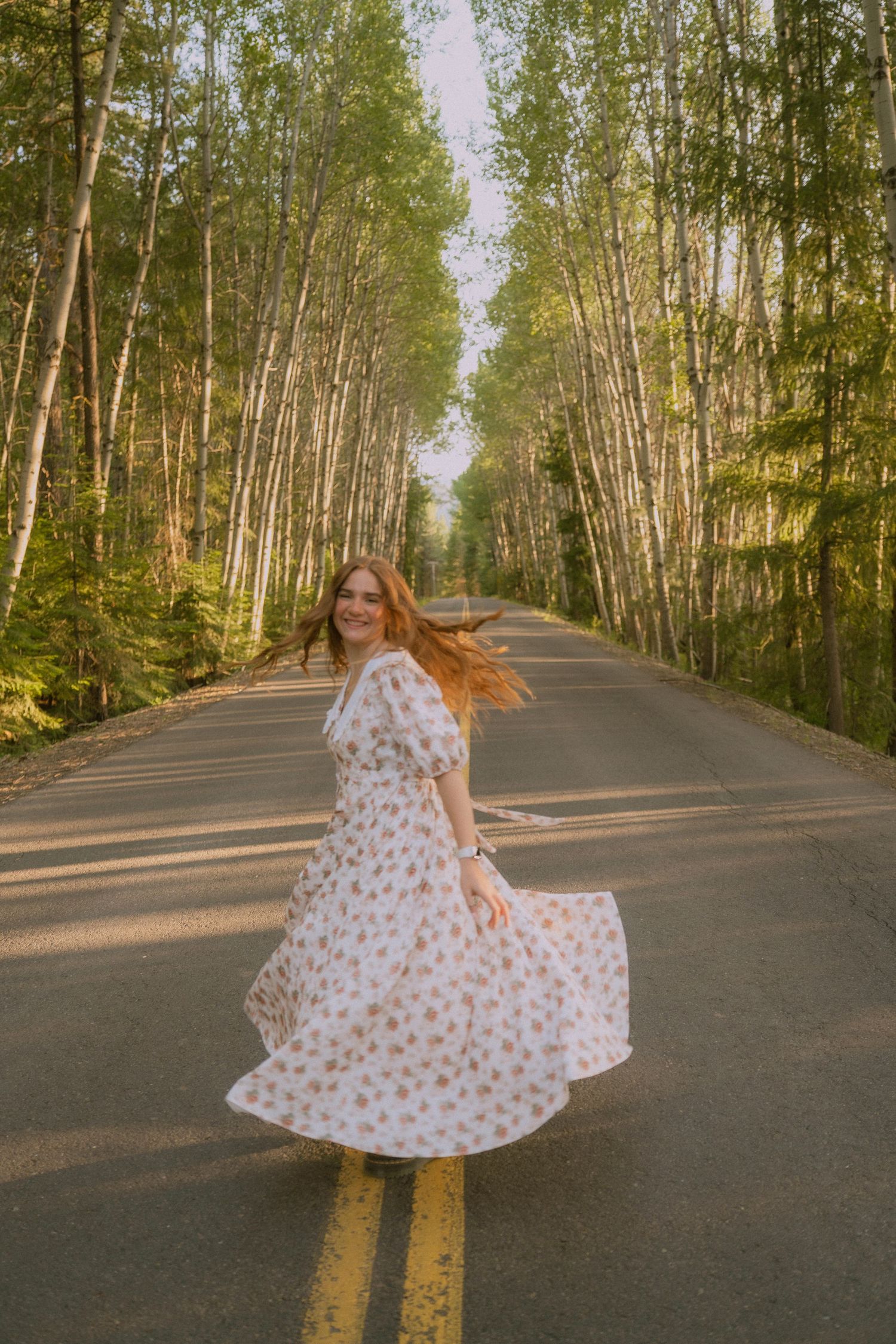 A floral dress twirls gracefully on a tree-lined road with golden sunlight filtering through birch trees.