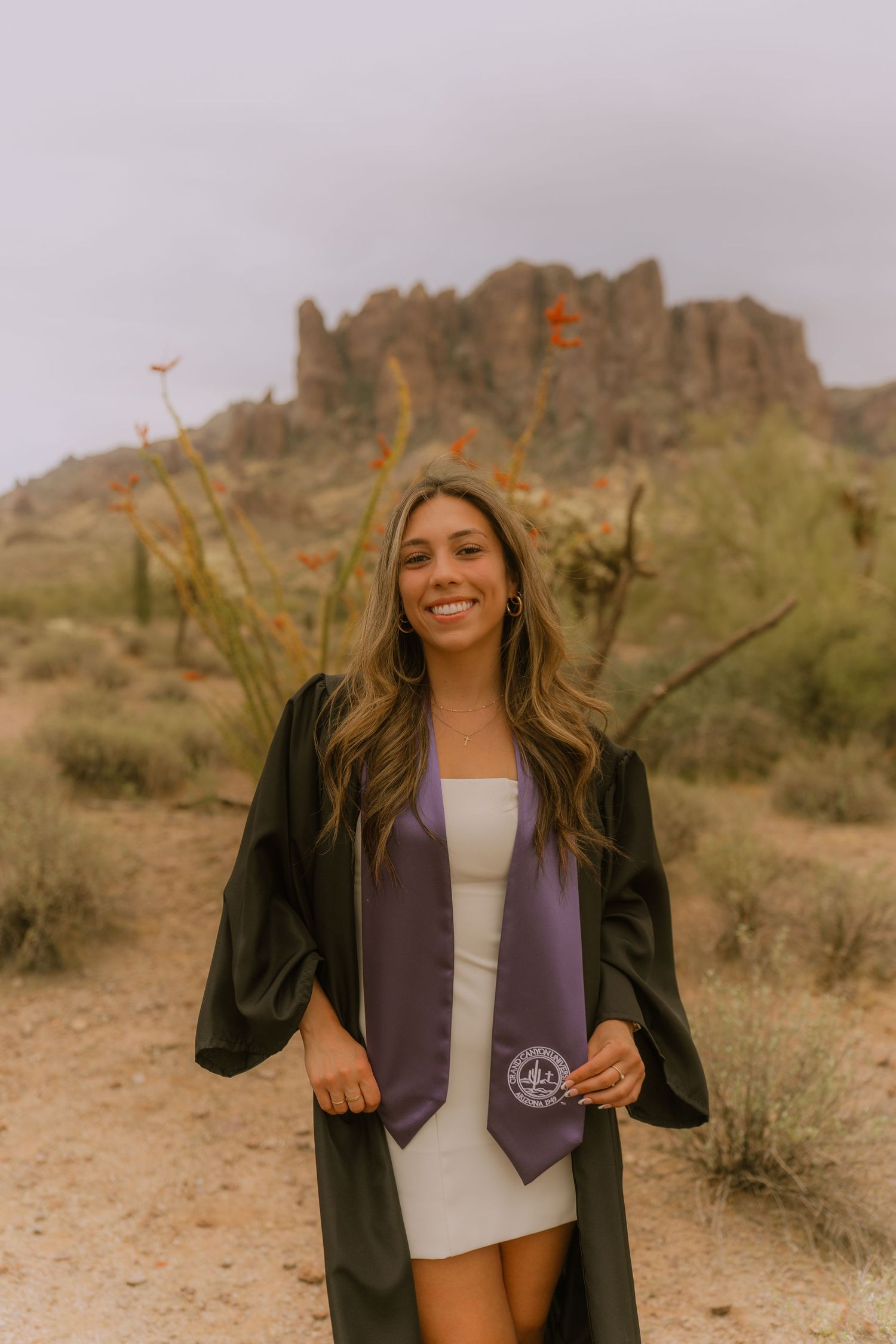 A graduate poses in cap and gown against a desert mountain backdrop at sunset with desert flora visible.