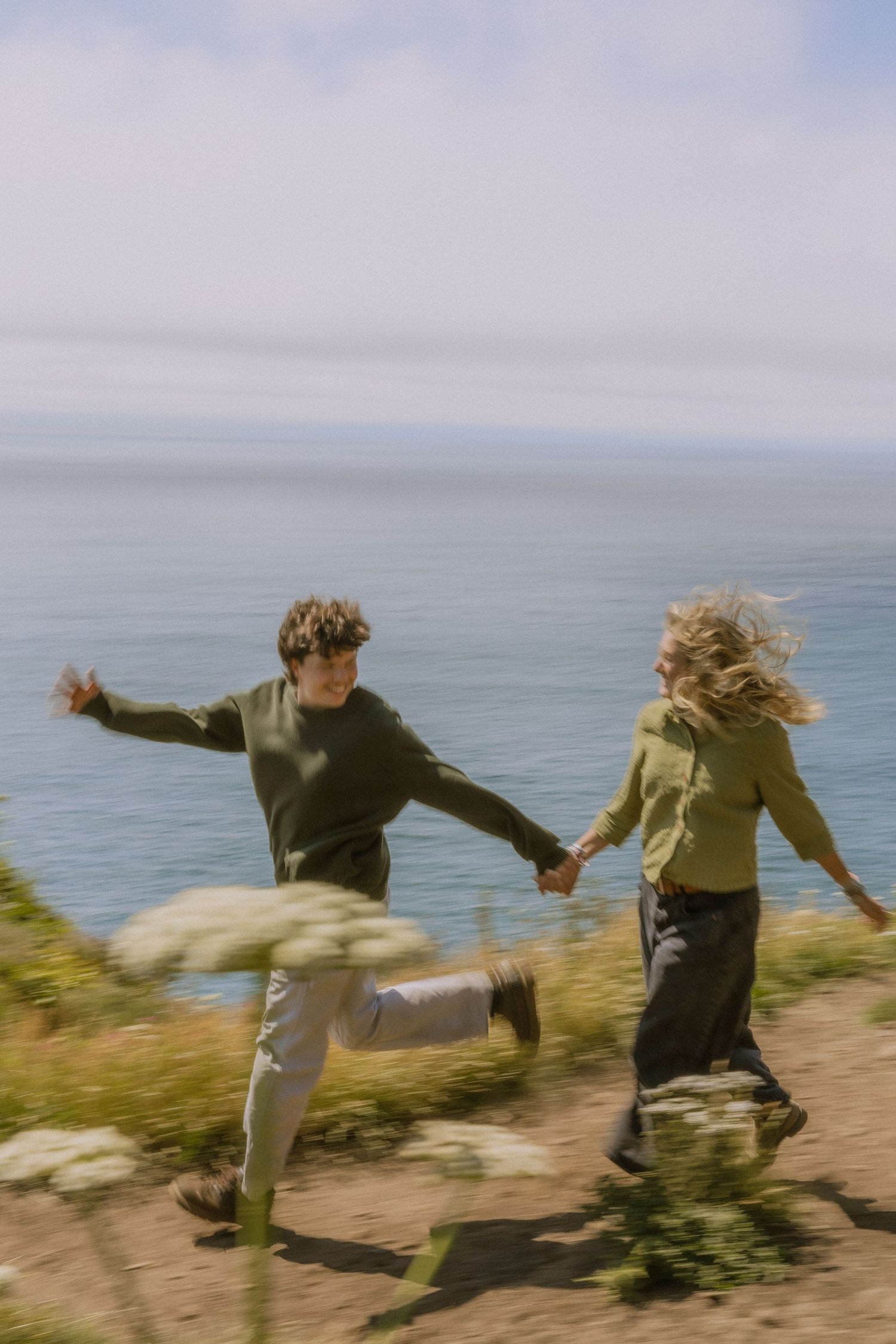 A sequence of motion showing two people playfully dancing and jumping on a coastal cliff overlooking the ocean.