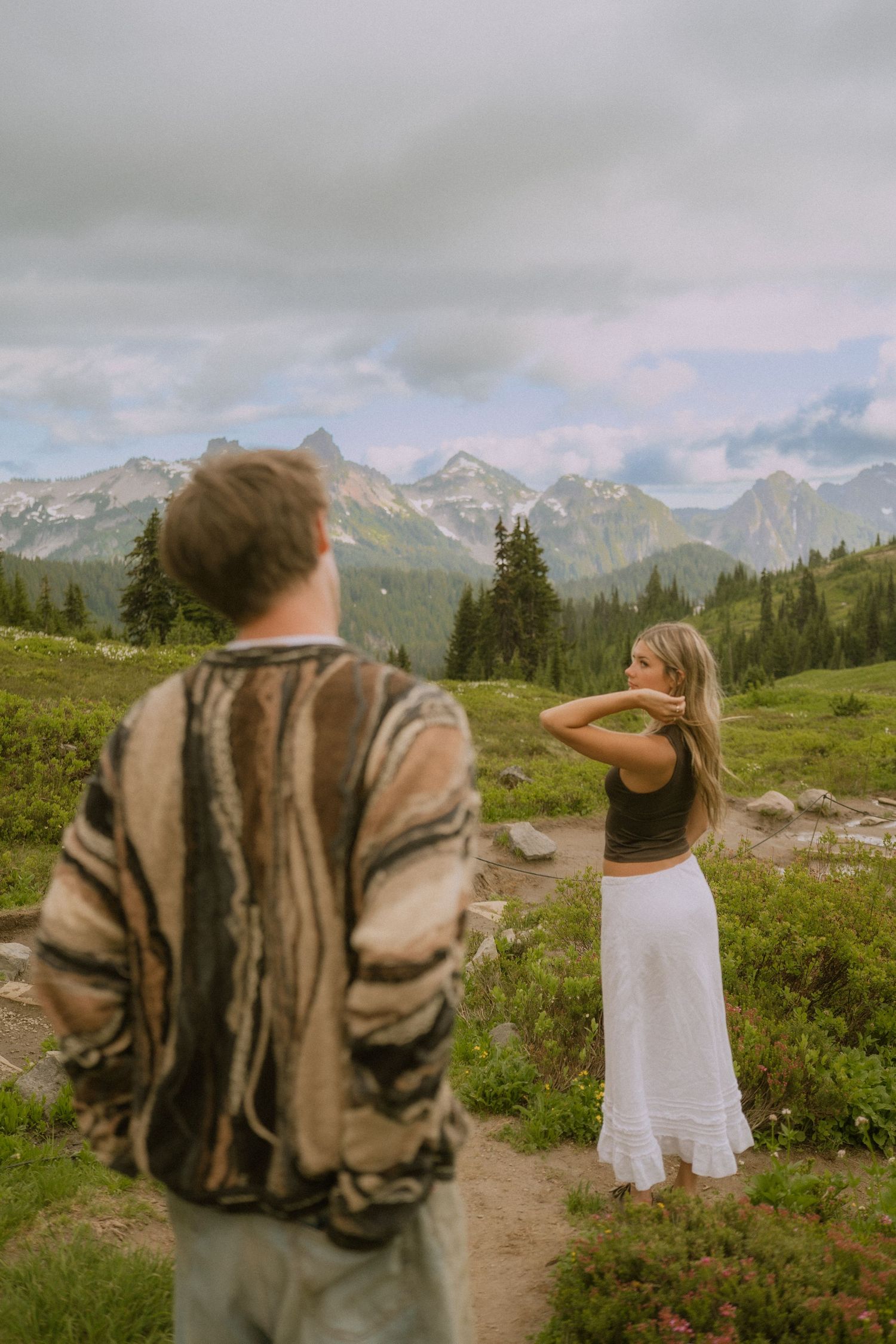 Two people stand on a mountain trail surrounded by alpine meadows and distant snow-capped peaks.