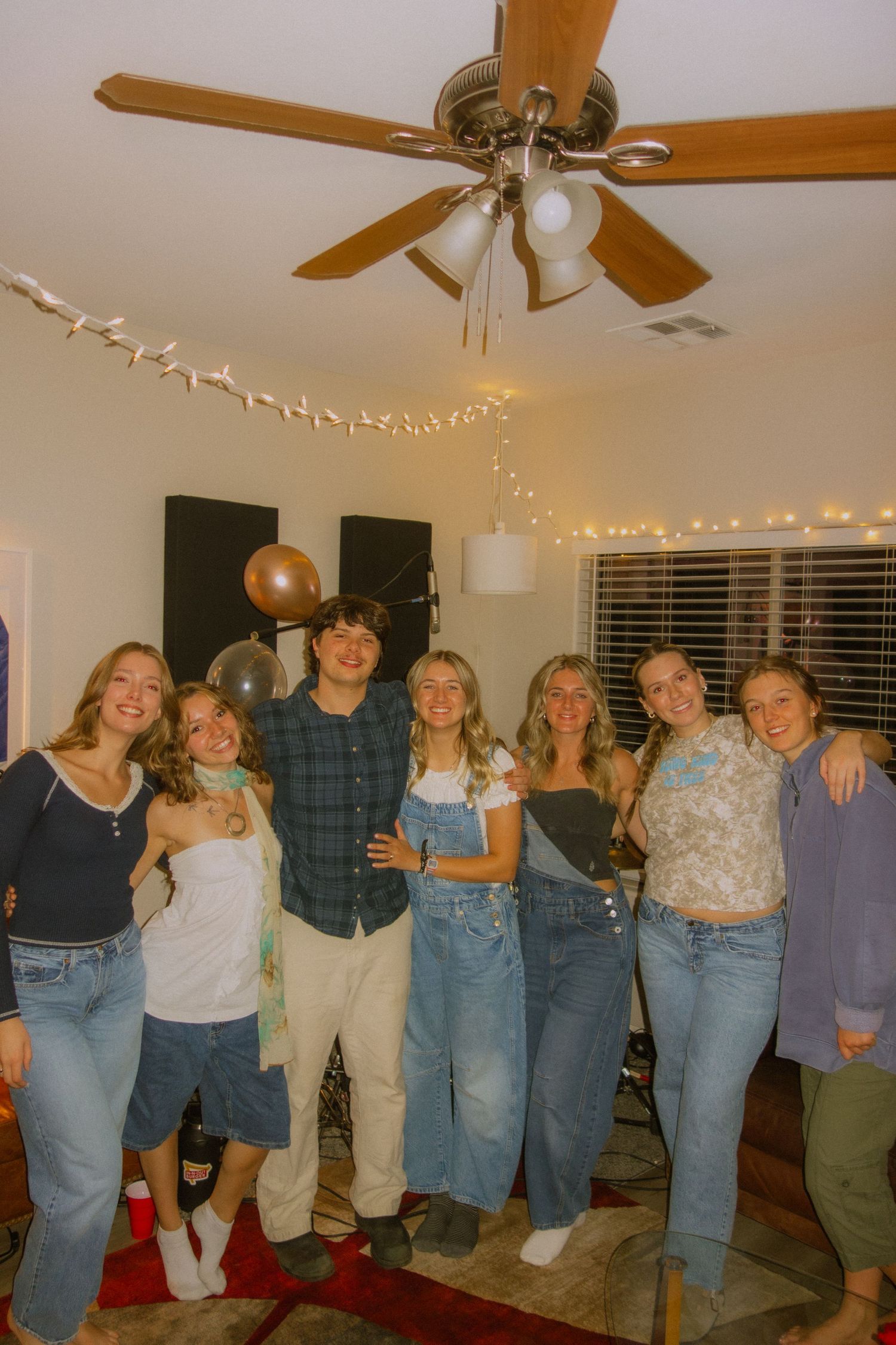 Group of friends gathered at a house party under string lights and ceiling fan in casual denim outfits.