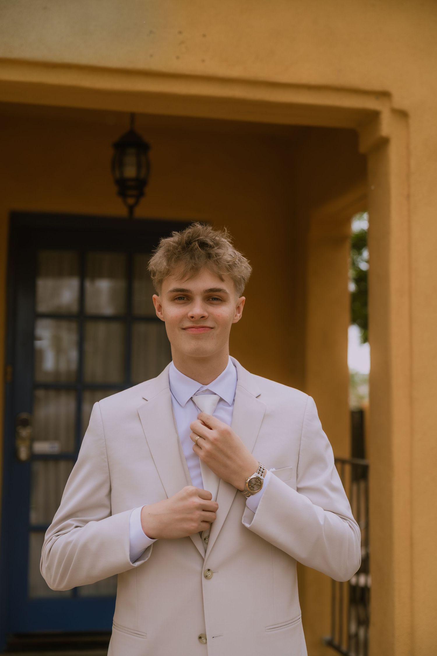 A person in a white suit adjusts their tie while standing in front of a beige building with arched doorways.