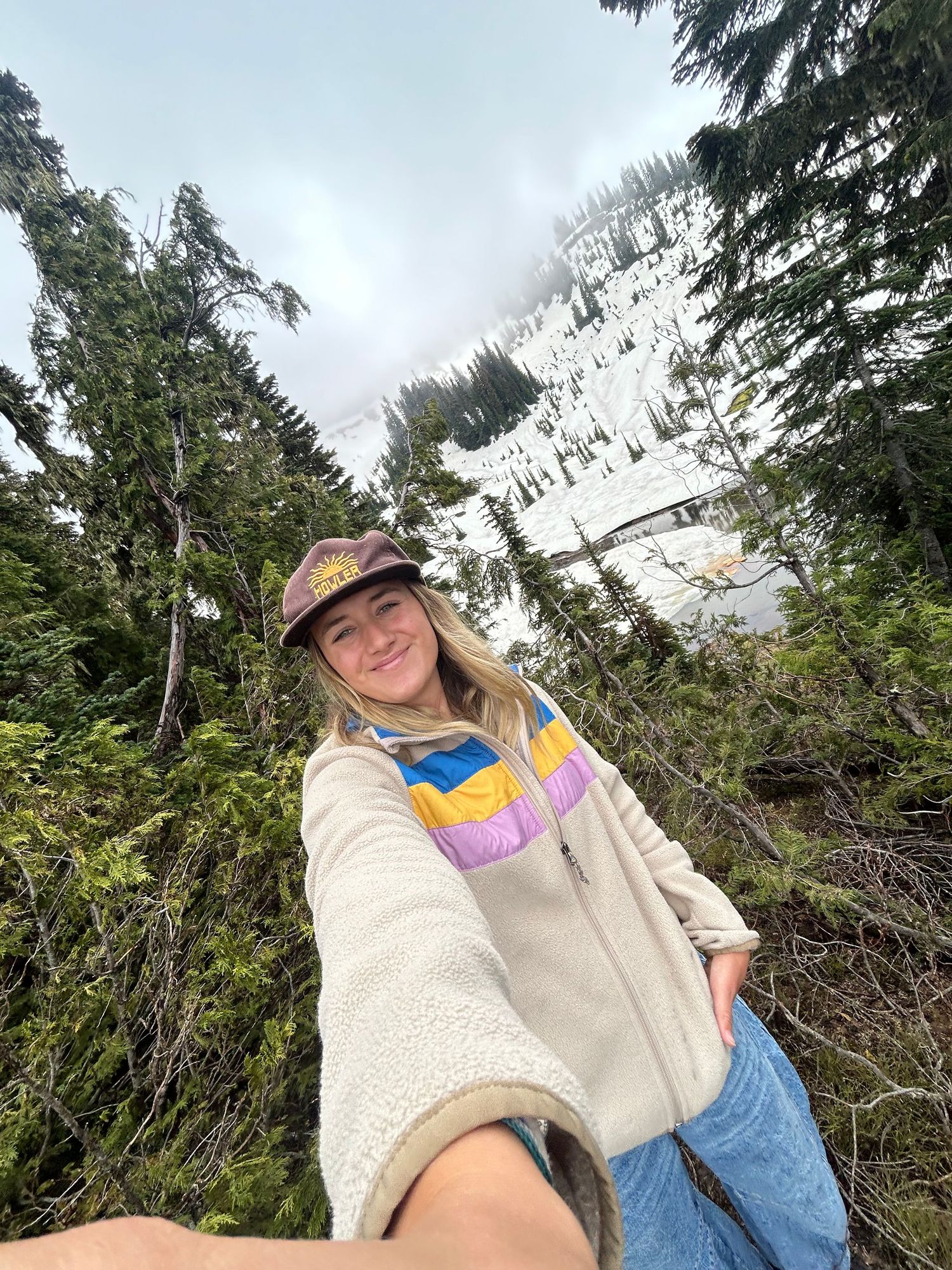 A hiker in casual attire and baseball cap takes selfies against a backdrop of evergreen trees and mountain scenery.