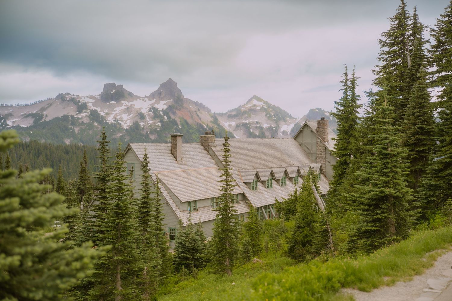 A scenic mountain lodge nestled among evergreen trees with snow-capped peaks visible in the misty background.