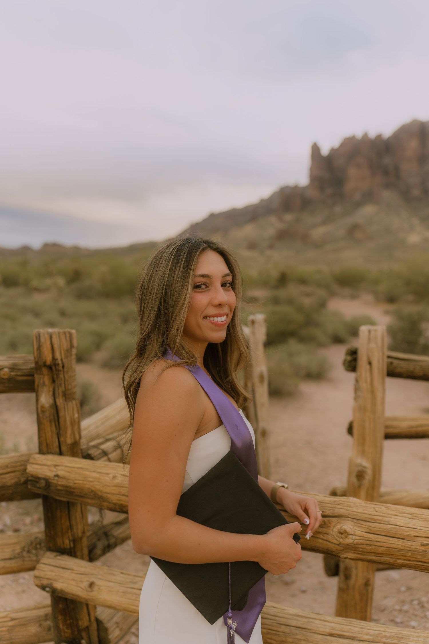 A person poses in a black and purple outfit against a desert landscape with wooden fencing and mountains in the background.