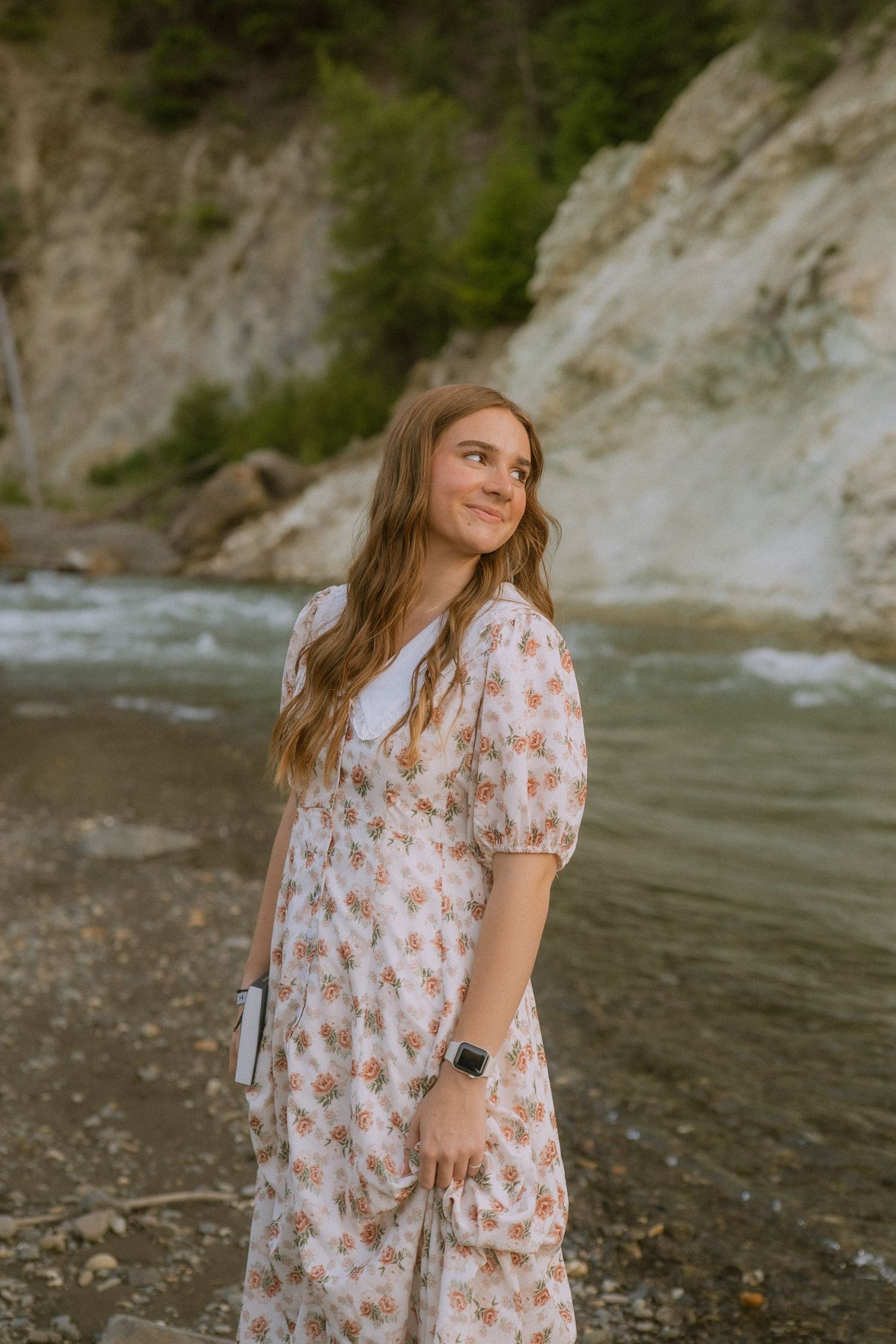 A flowing floral dress catches the light along a rocky riverbank with cliffs and rushing water.