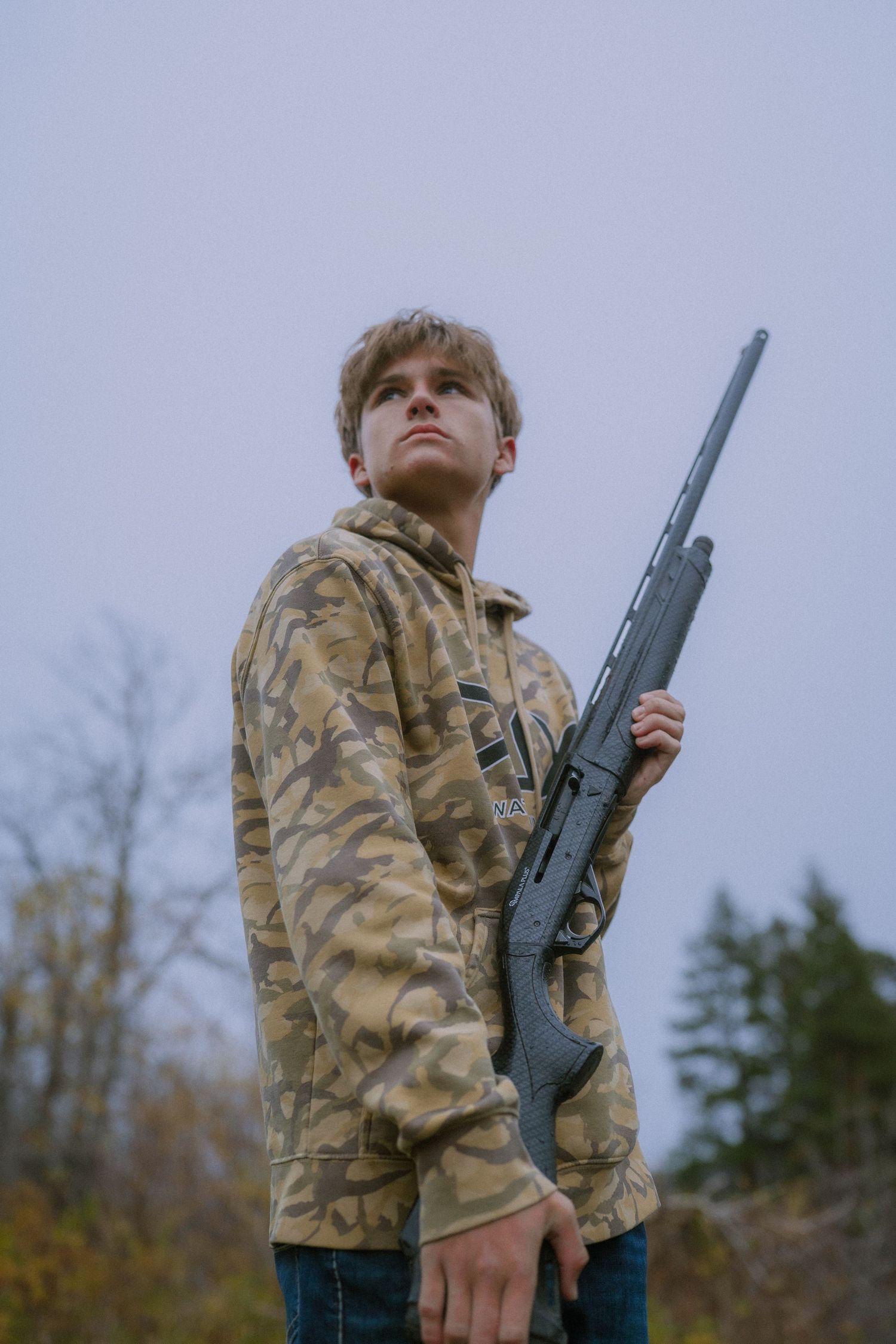 Person in camouflage jacket holds a rifle while looking up at overcast sky in outdoor setting.